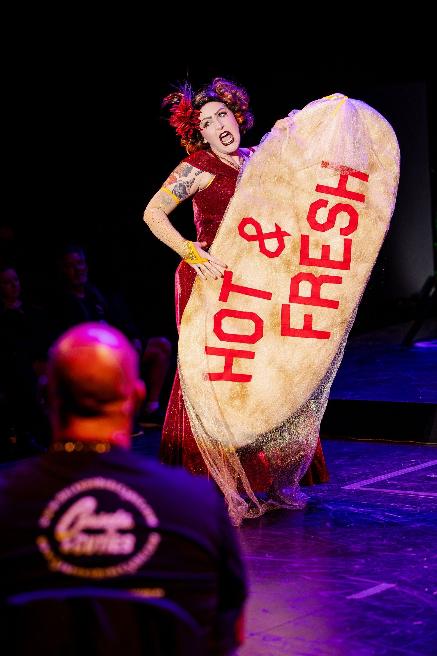 A burlesque performer in red holds a large prop reading Hot and Fresh on stage at a live cabaret show.