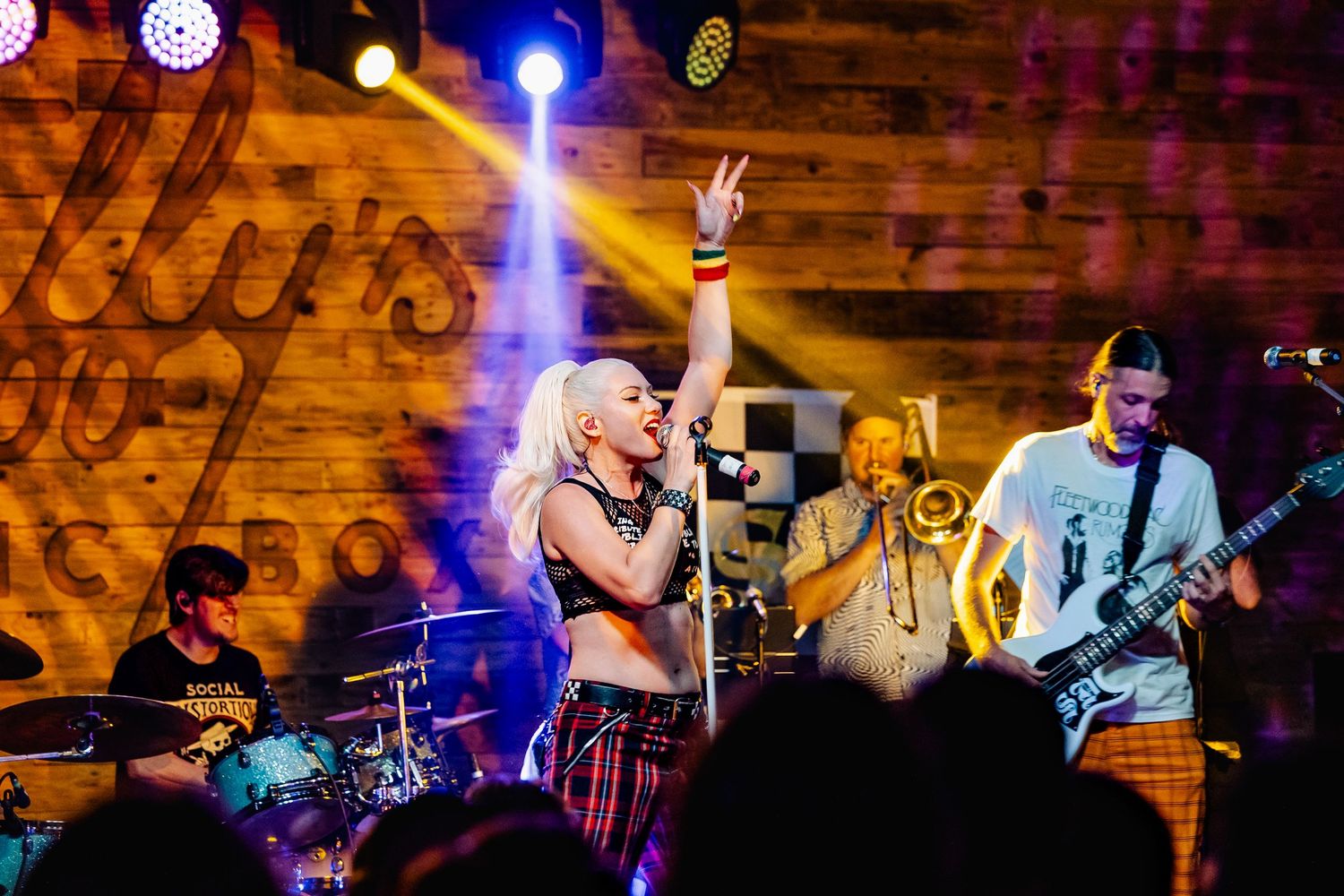 Blonde female singer performs on stage with band, making peace sign under yellow stage lights at a rock venue.