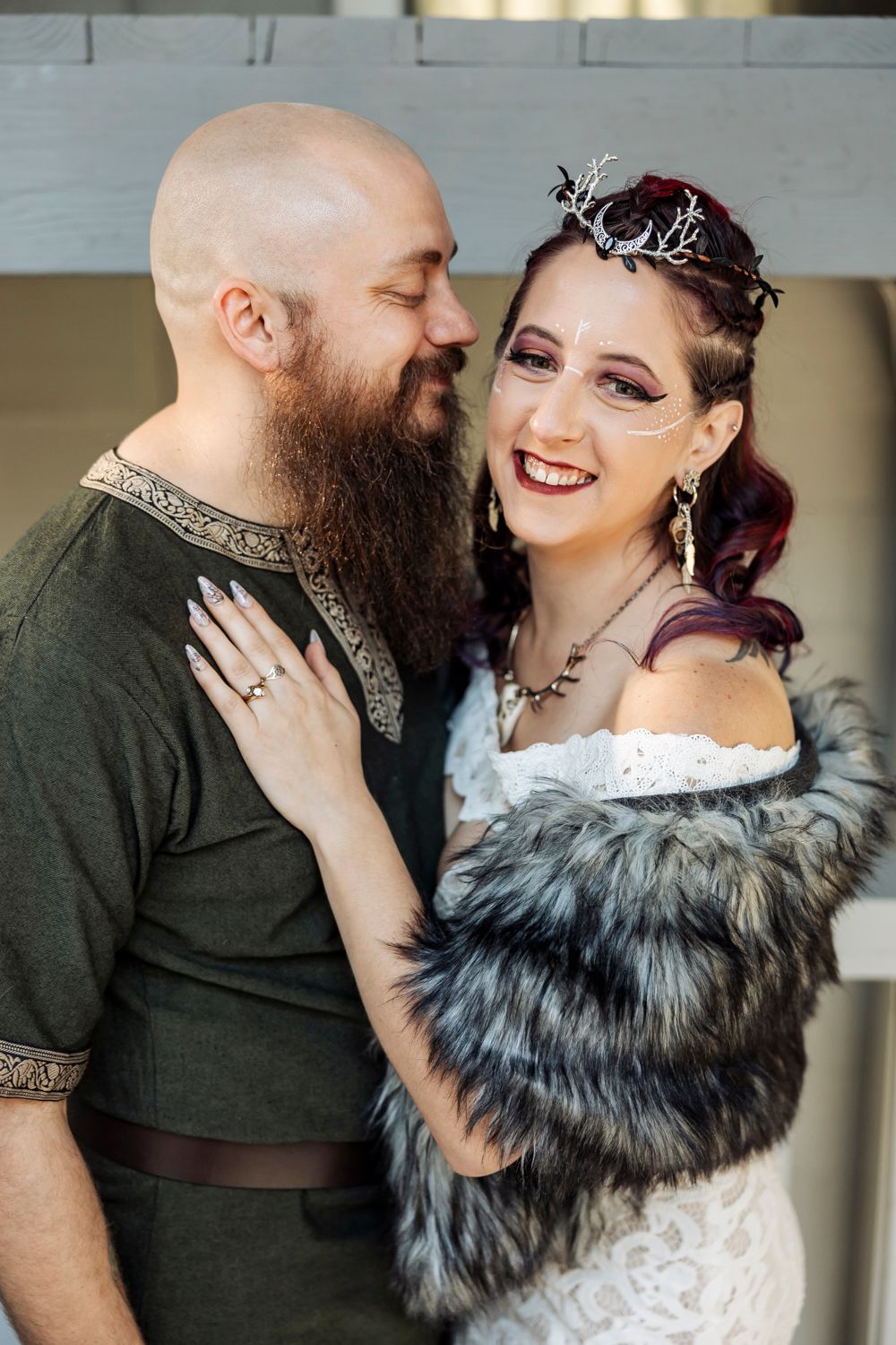 A bearded groom in medieval attire smiles at his bride wearing a floral crown and fur wrap at a fantasy wedding.