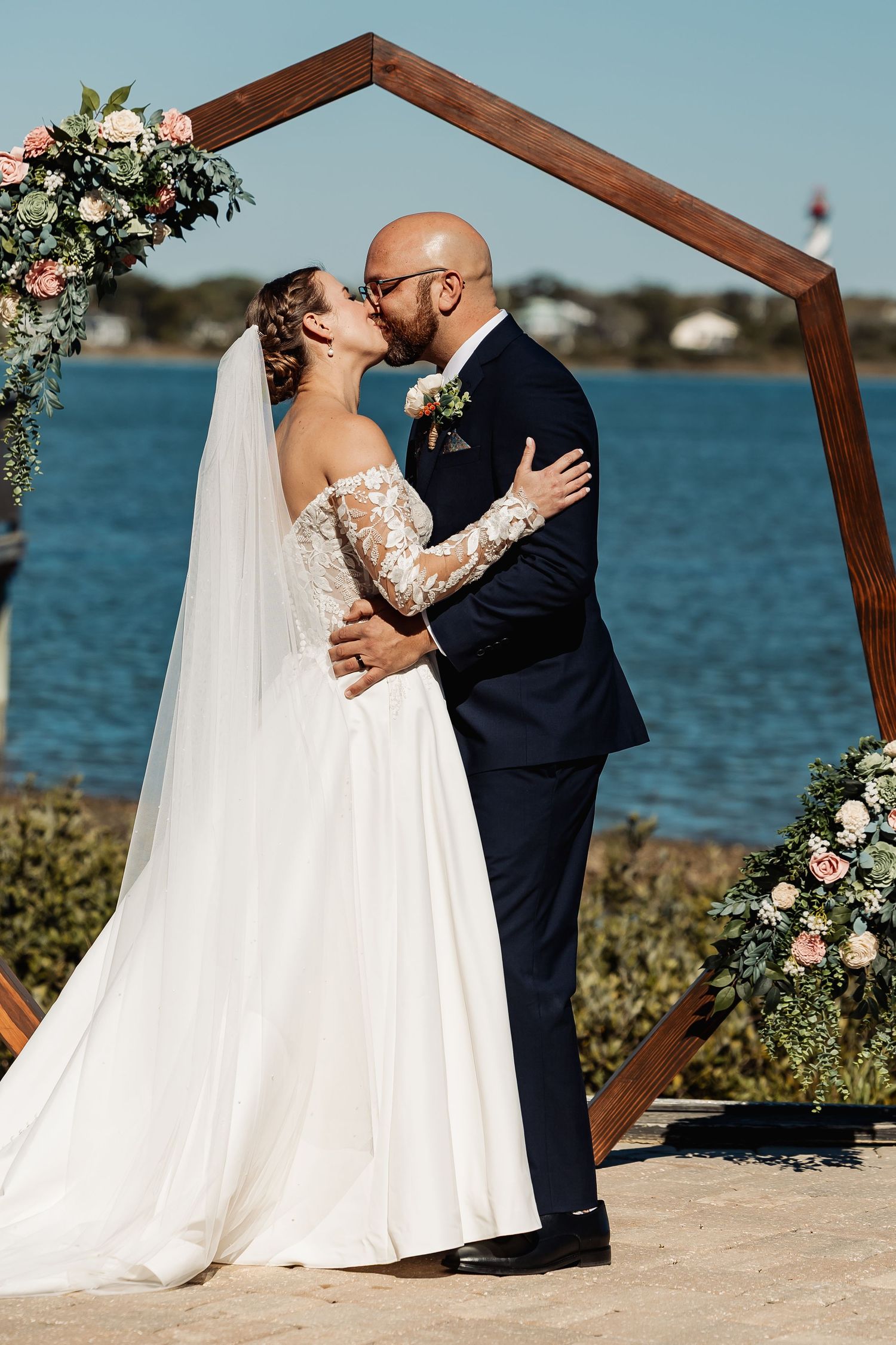 Bride and groom share first kiss under hexagonal wooden arch adorned with pink florals by waterfront.
