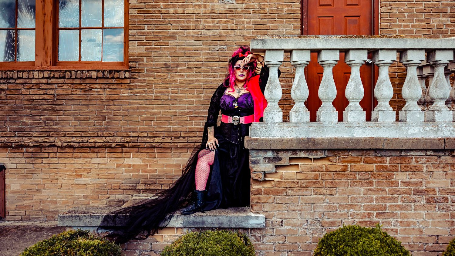 Woman in black gothic dress with purple corset poses confidently on stone steps by brick building.