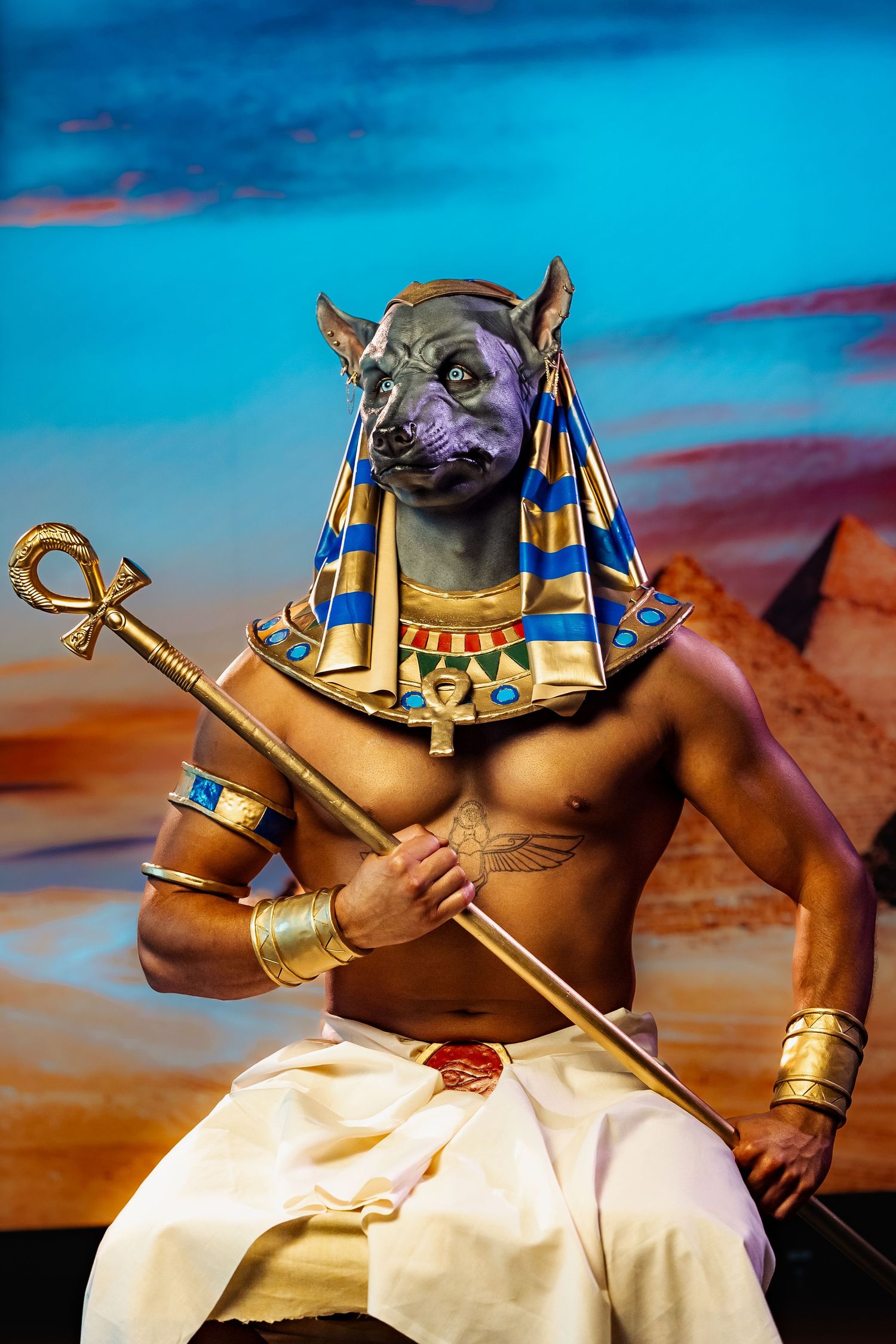 Person in Egyptian god Anubis costume with jackal mask holding staff against pyramid backdrop.
