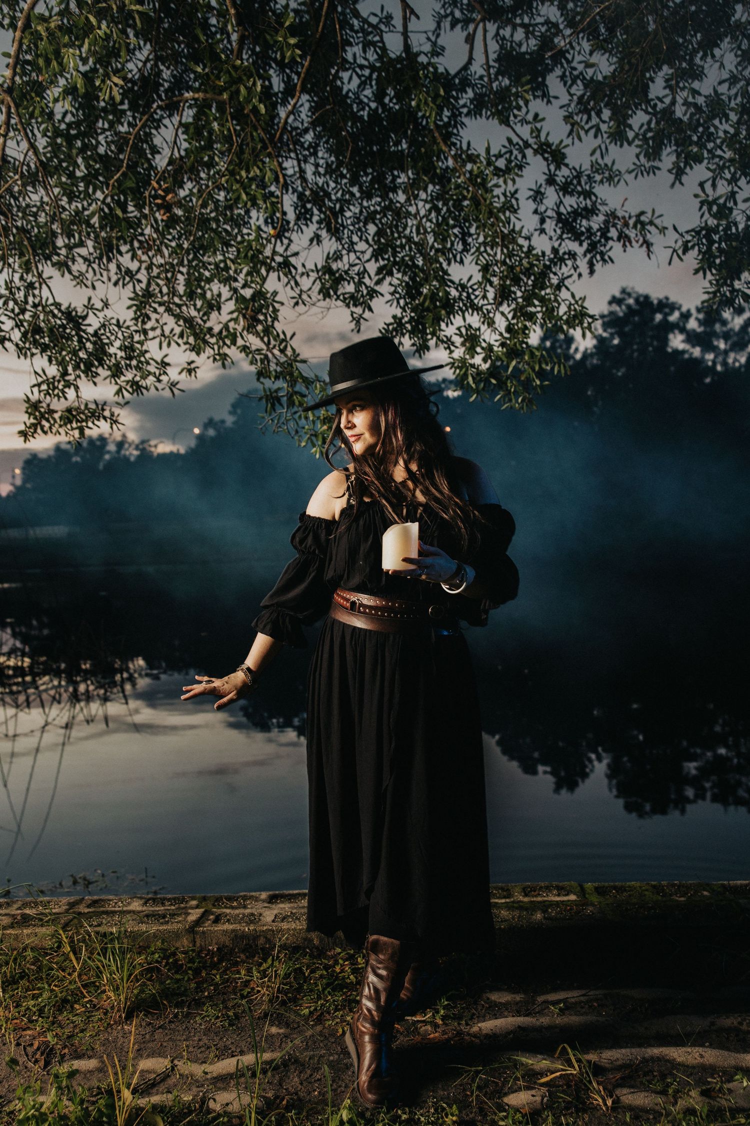 Woman in black witch costume holding a candle stands by a misty lake at dusk under willow branches.