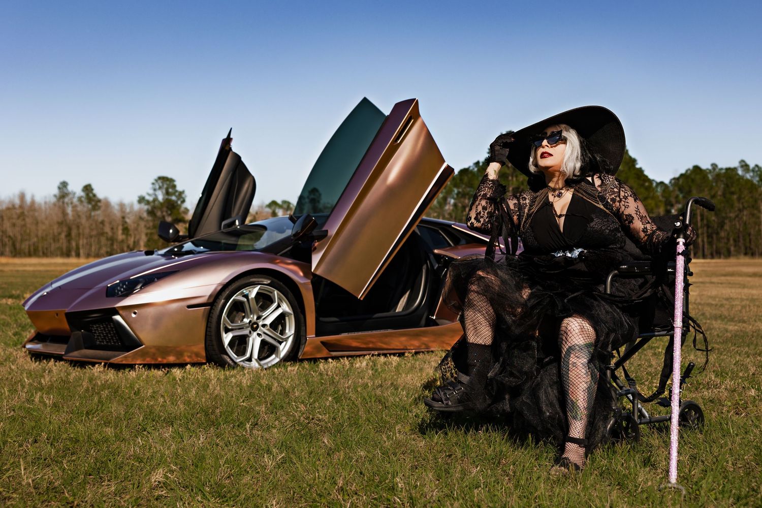 Woman in black gothic outfit and wide-brim hat poses beside a bronze Lamborghini Aventador with scissor doors open in a field.