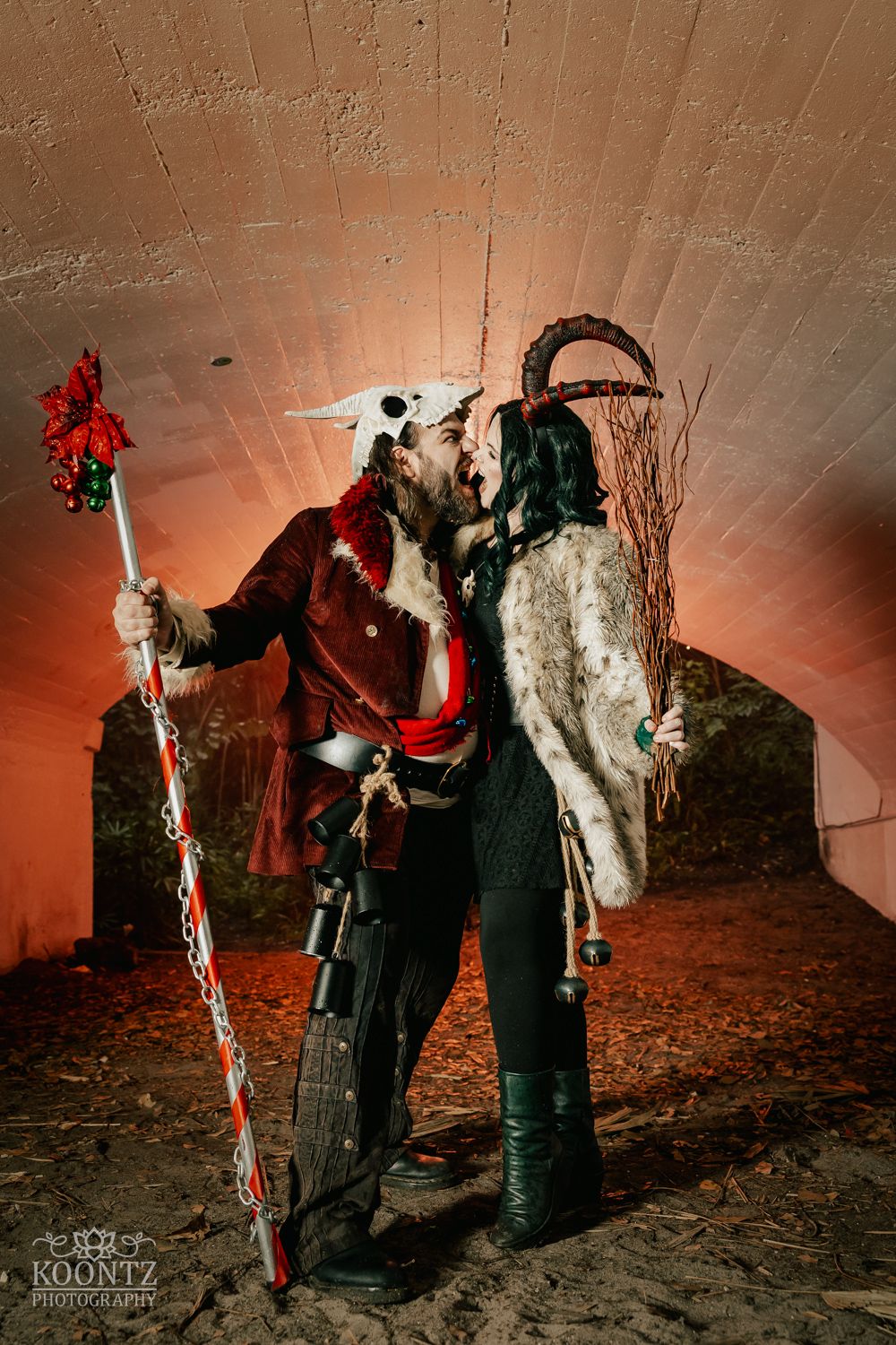 Couple in dark fantasy Krampus-inspired costumes share a kiss in a dramatically lit tunnel setting.