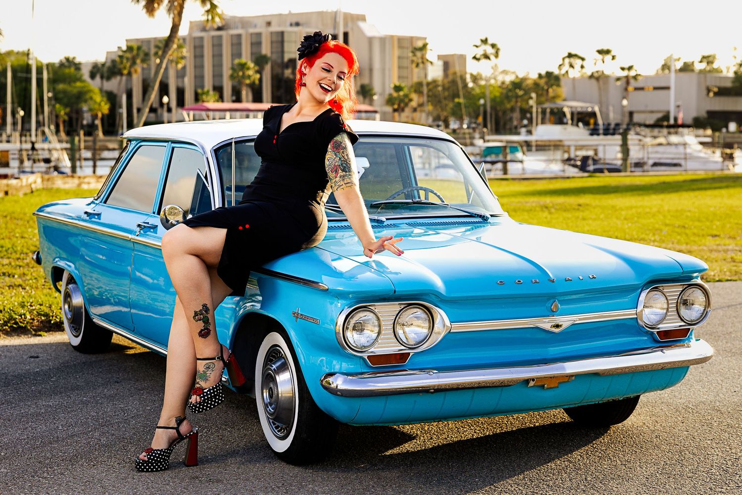 Tattooed redhead pinup model sitting on blue classic car hood in black dress.