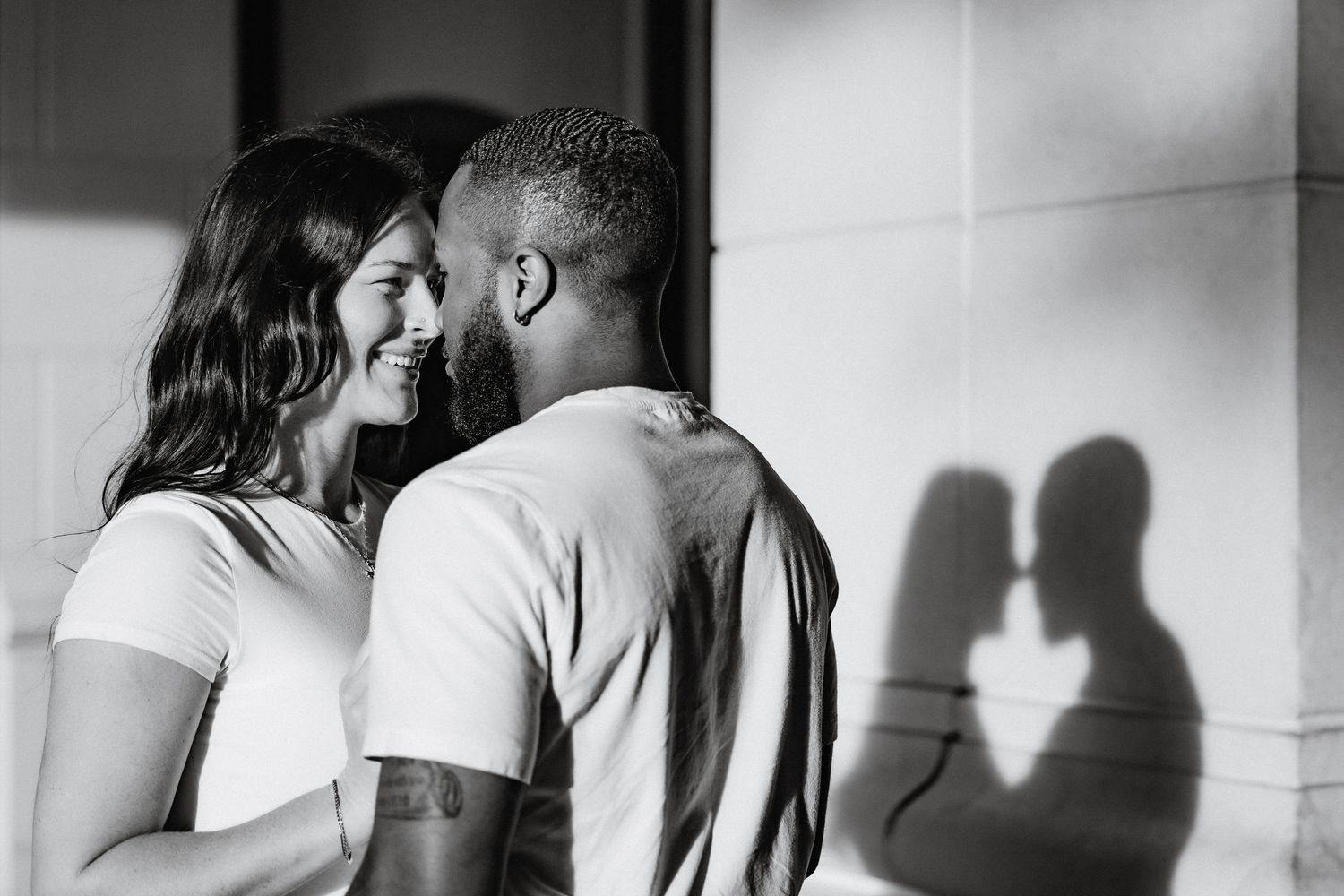 Black and white photo of couple smiling at each other with romantic shadow cast on sunlit wall.