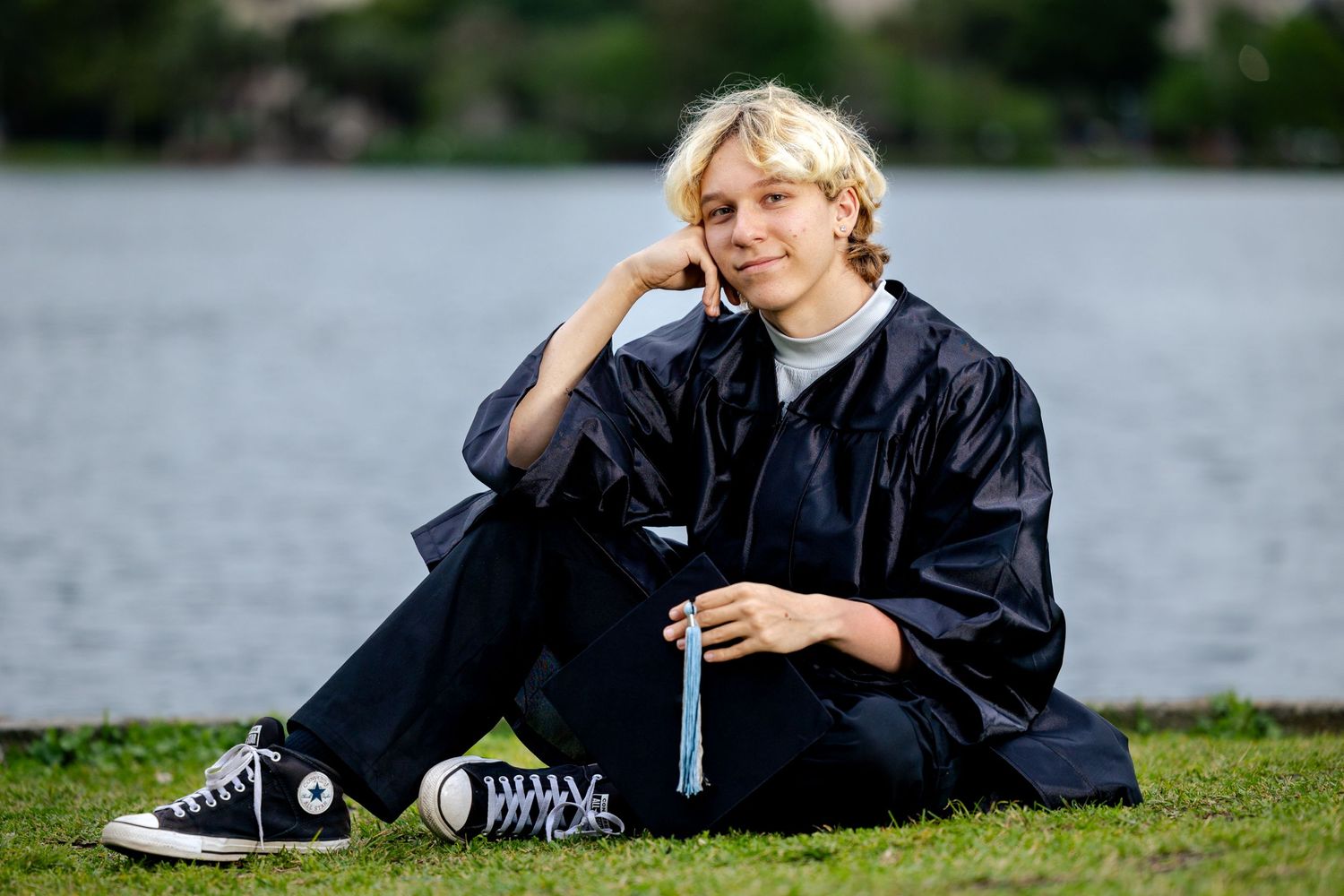 Person in graduation gown relaxes on grass by lake wearing black Converse sneakers.
