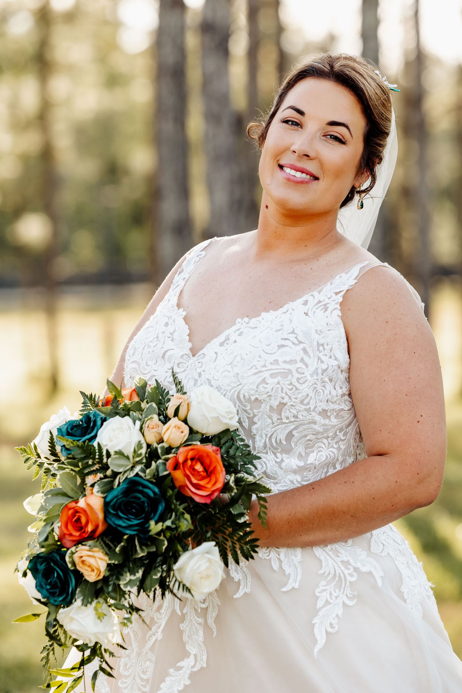 Beautiful bride holding vibrant orange and blue bouquet against pine forest backdrop.