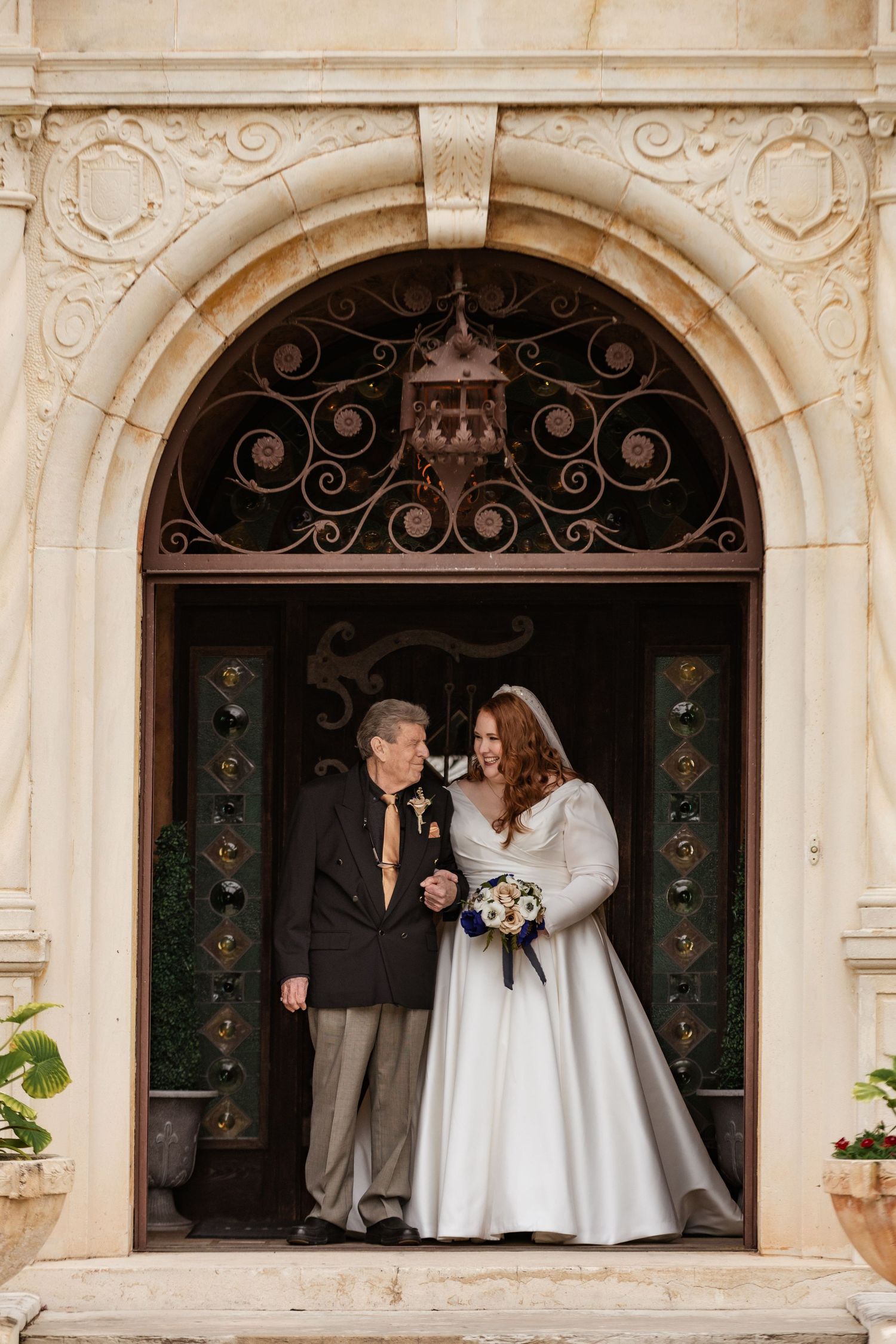 Bride in a white ball gown holding a bouquet standing with her father under an ornate stone archway.
