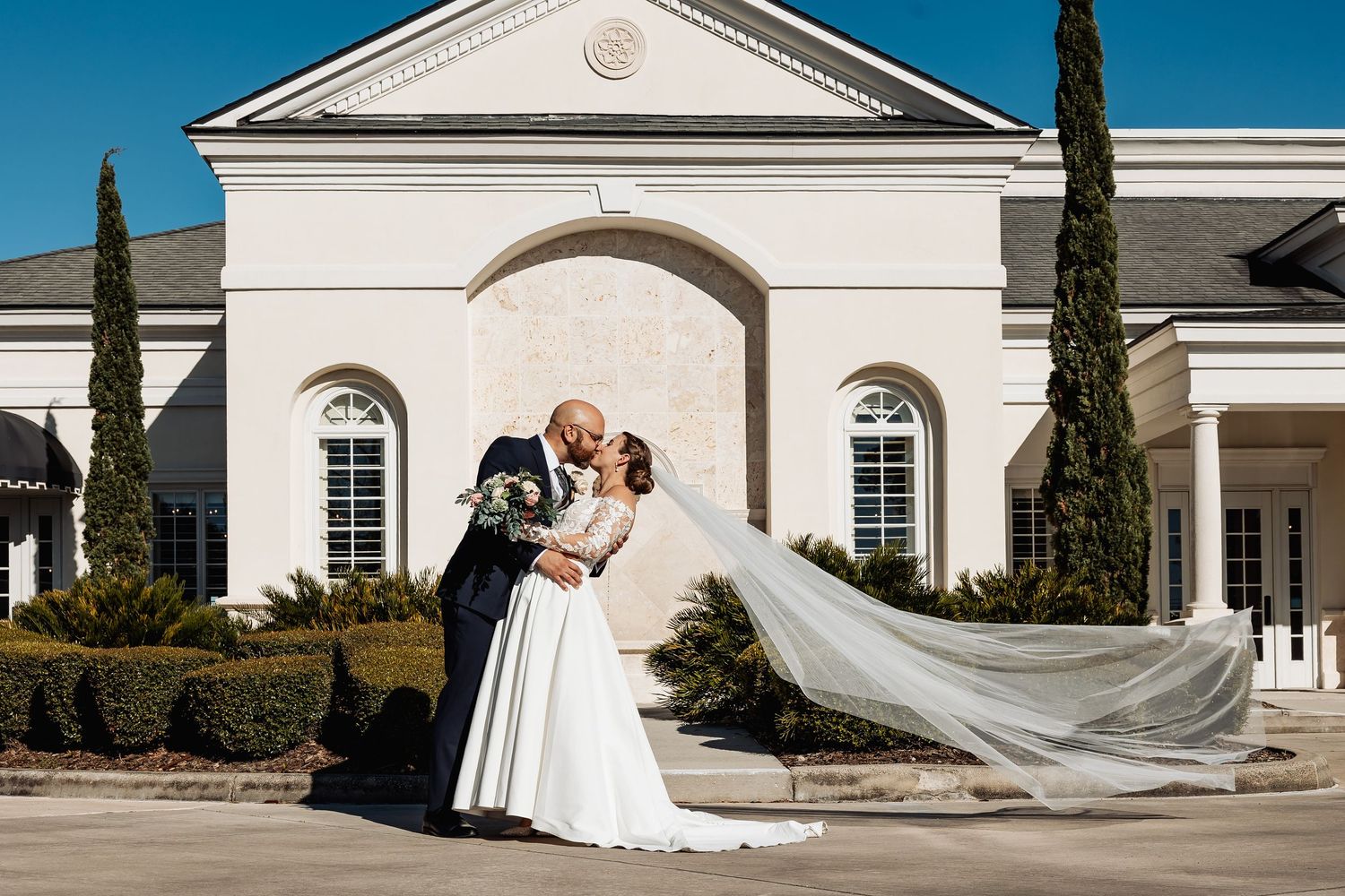 Bride and groom kissing outside elegant white venue with long veil flowing in wind.