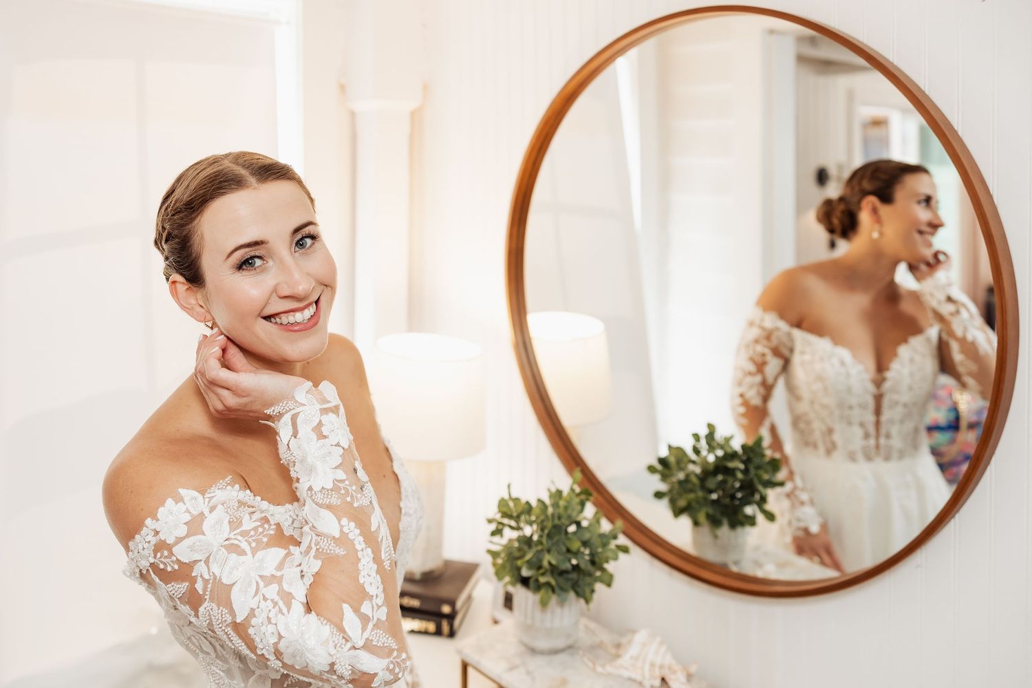 Smiling bride in lace off-shoulder wedding dress admiring her reflection in a round mirror.