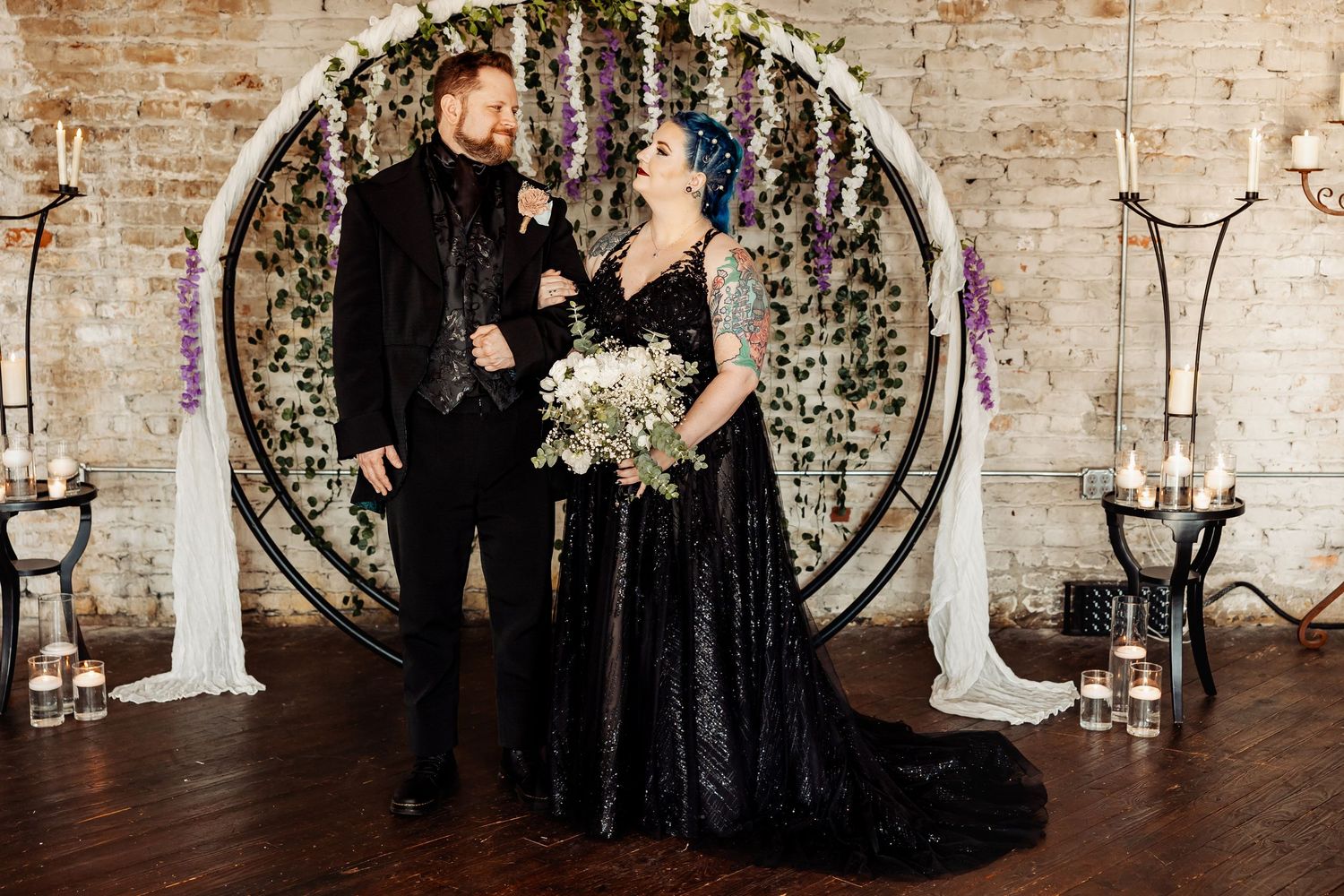Bride in black sequin gown and groom in black suit stand before floral circle arch with purple wisteria.