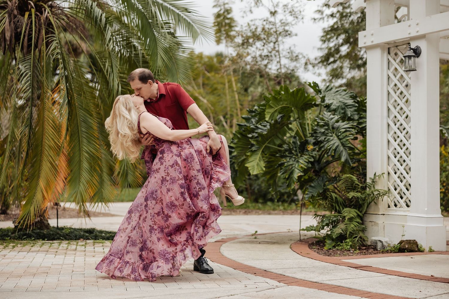Couple sharing a passionate dip kiss outdoors near a white lattice gazebo with palm trees.