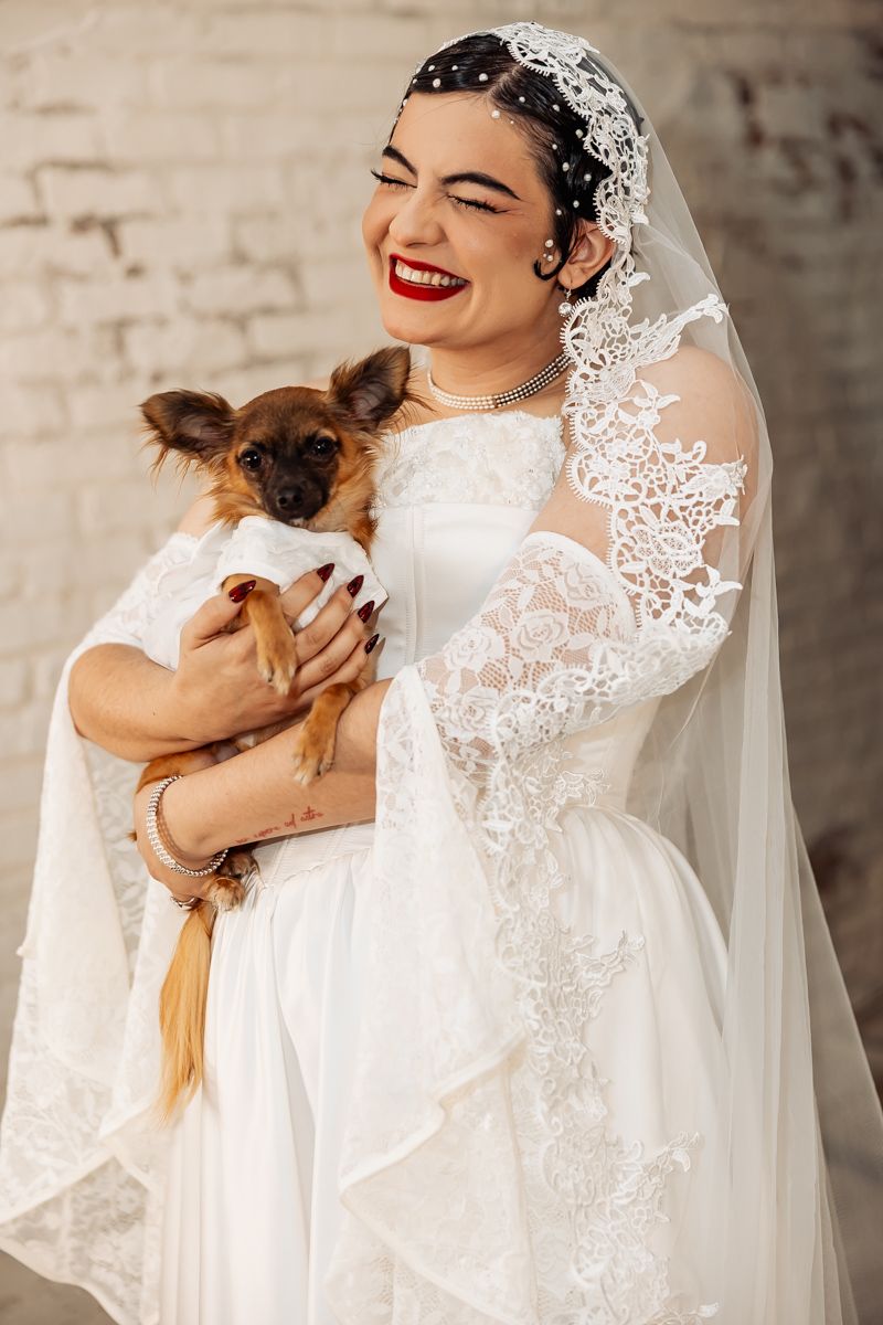 Smiling bride in lace wedding dress and veil holds small brown dog dressed in white at wedding.