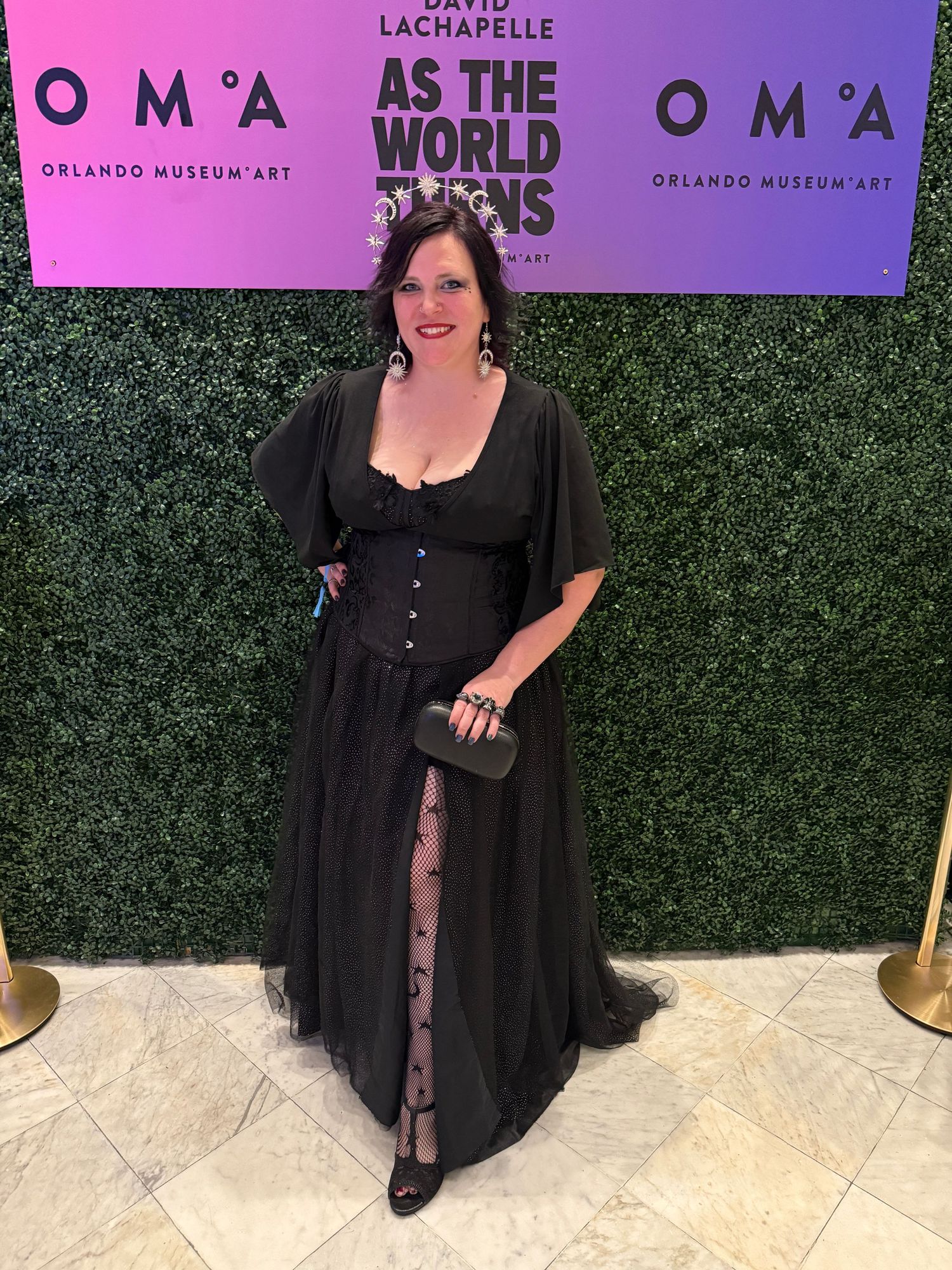 A person poses in a flowing black dress with buttons and sheer details at an event with a green hedge wall and pink signage.