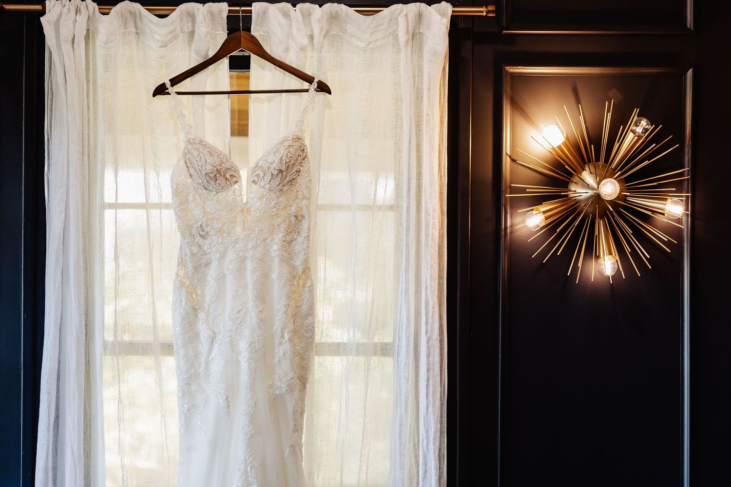 Lace bridal gown hanging on curtain rod beside a gold starburst wall sconce.