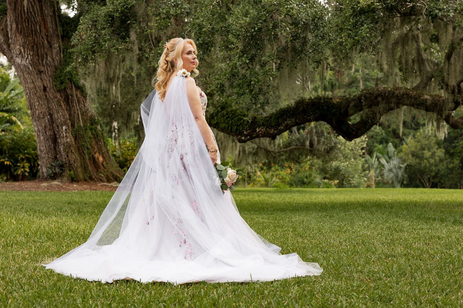 Bride in floral wedding gown with long veil standing beneath Spanish moss-draped oak tree.