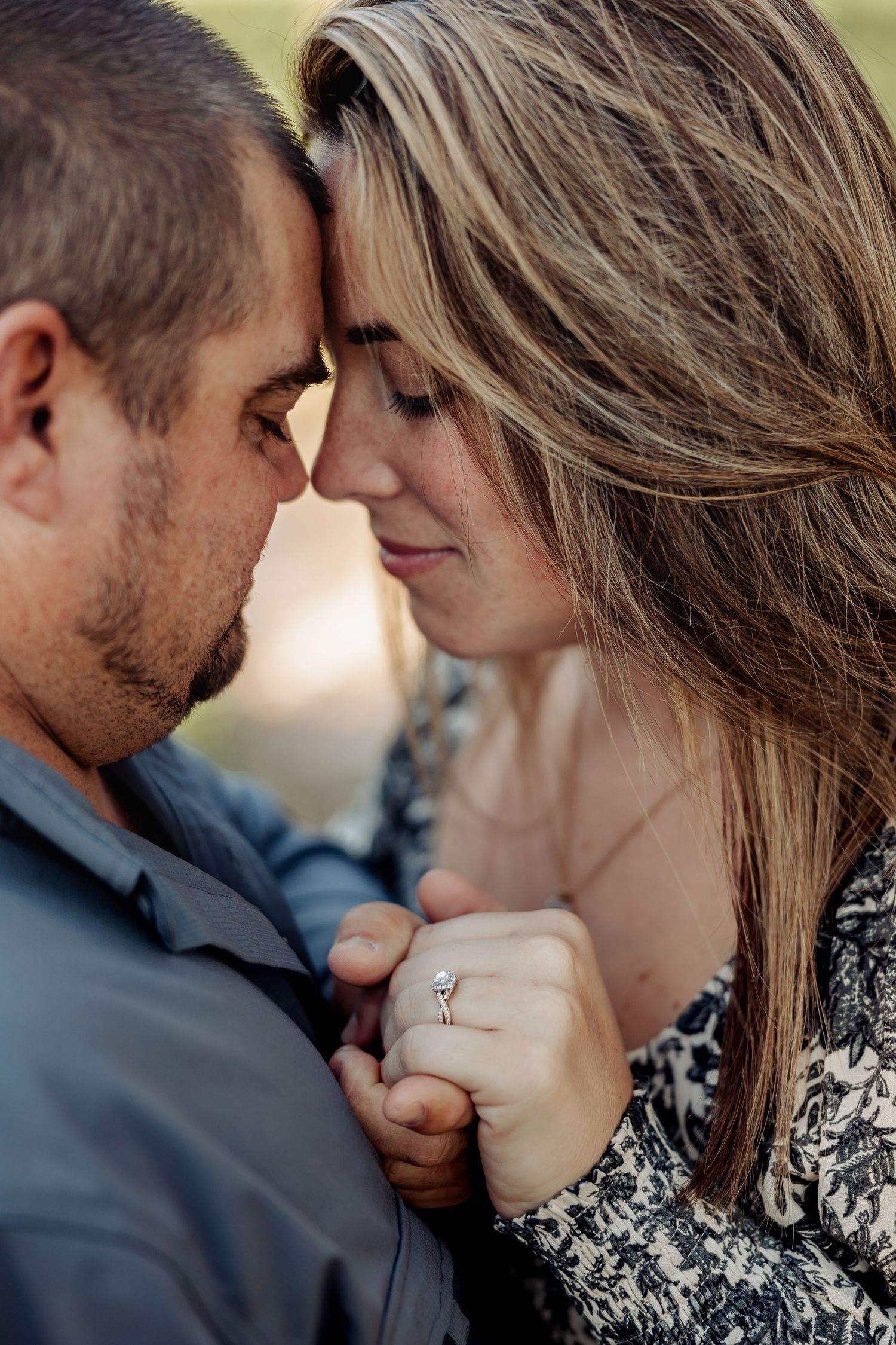 An engaged couple holds hands and touches foreheads in an intimate close-up showing a diamond ring.