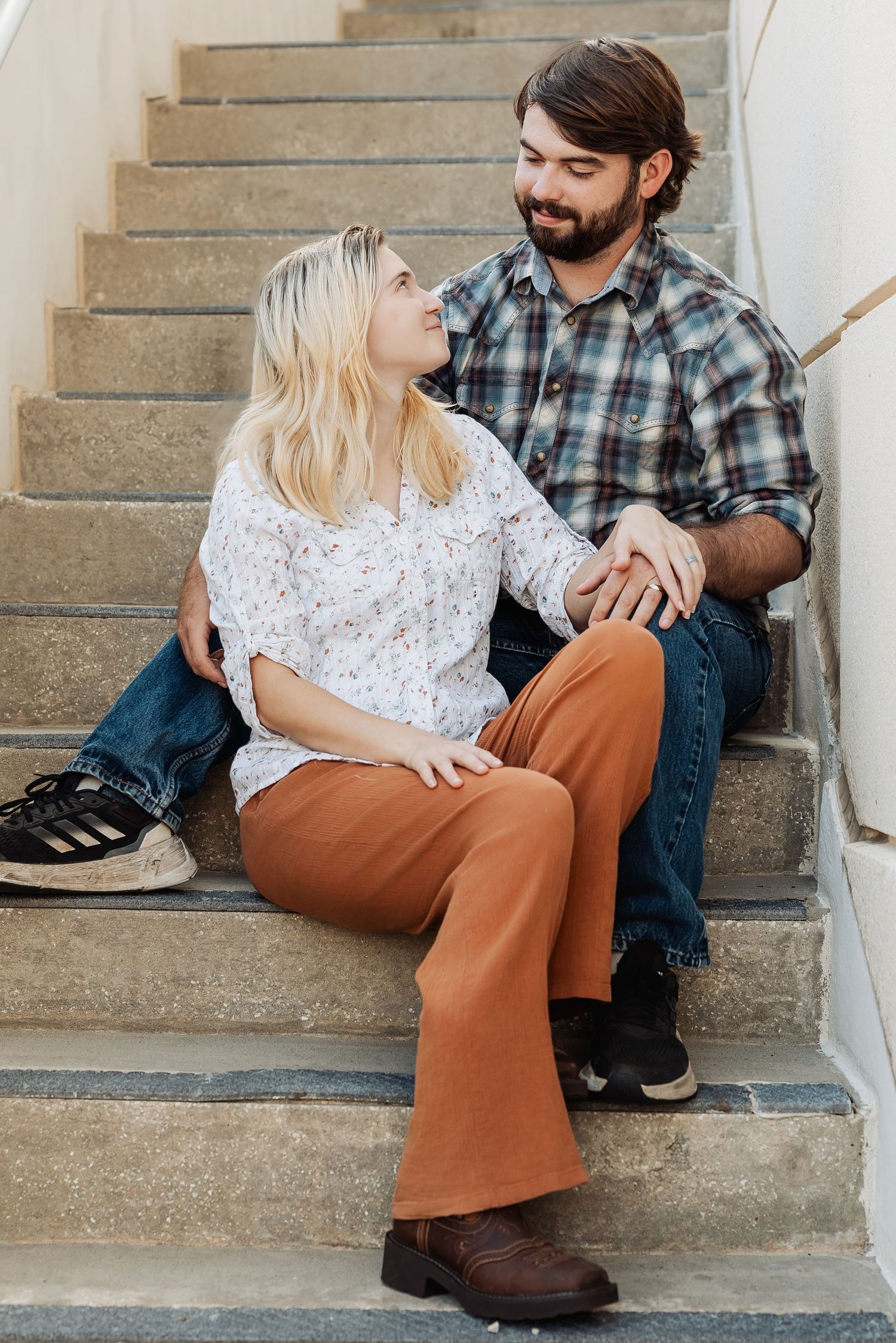 Young couple sitting on outdoor steps, woman in floral top and rust pants, man in plaid shirt.