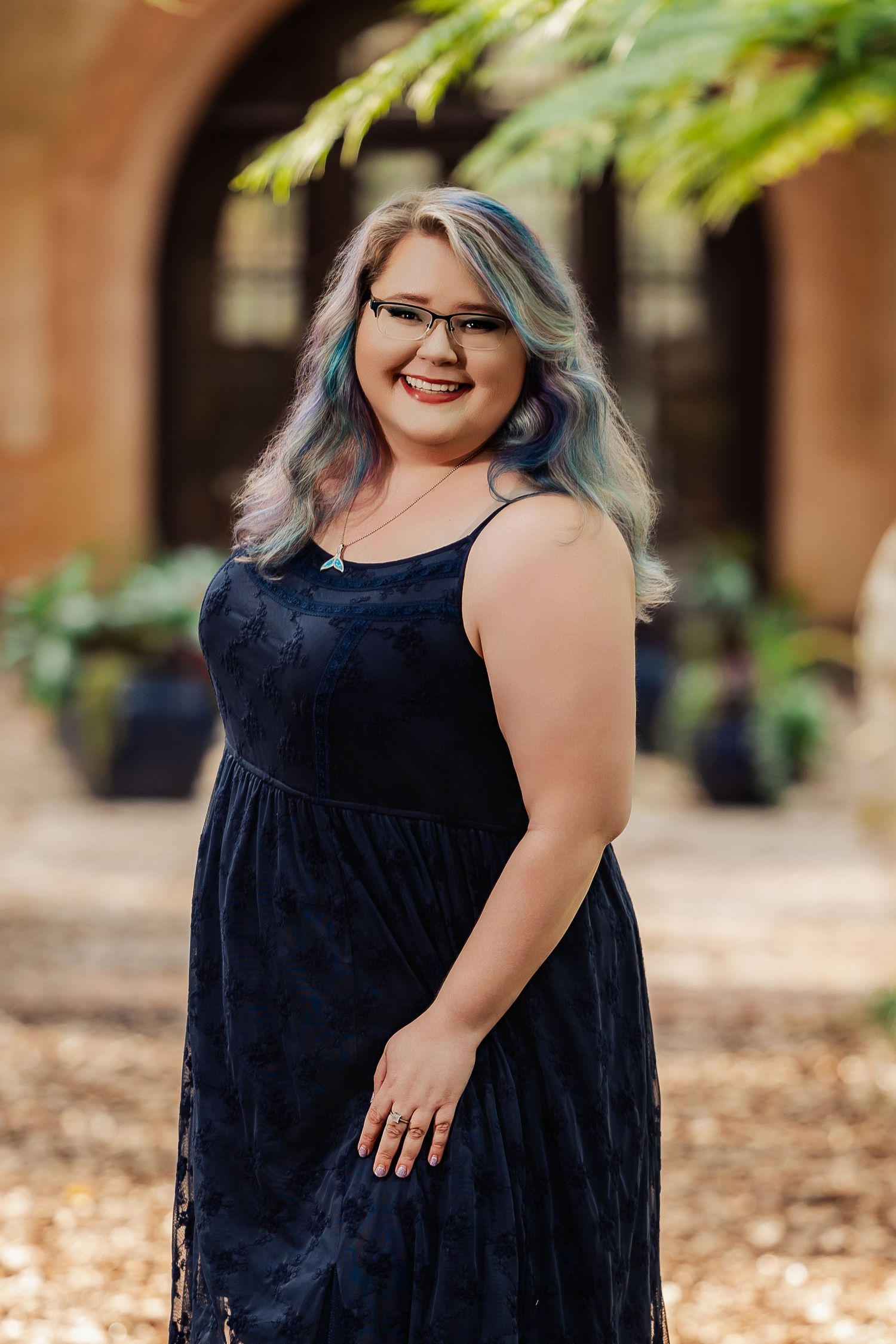 A smiling woman with colorful hair and glasses wearing a navy blue lace dress poses outdoors near an archway.