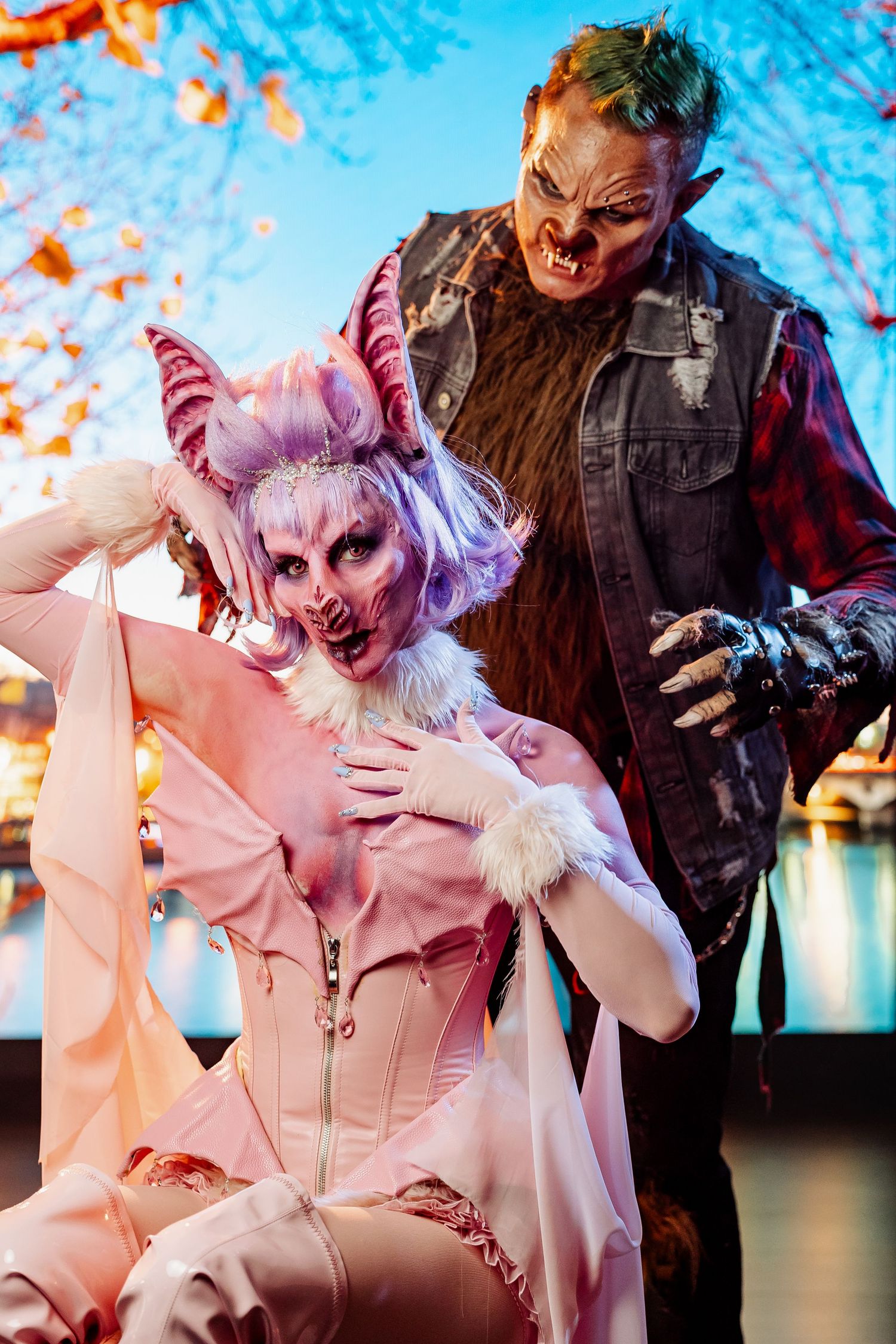 Two people in elaborate Halloween demon costumes pose together against a night cityscape backdrop.
