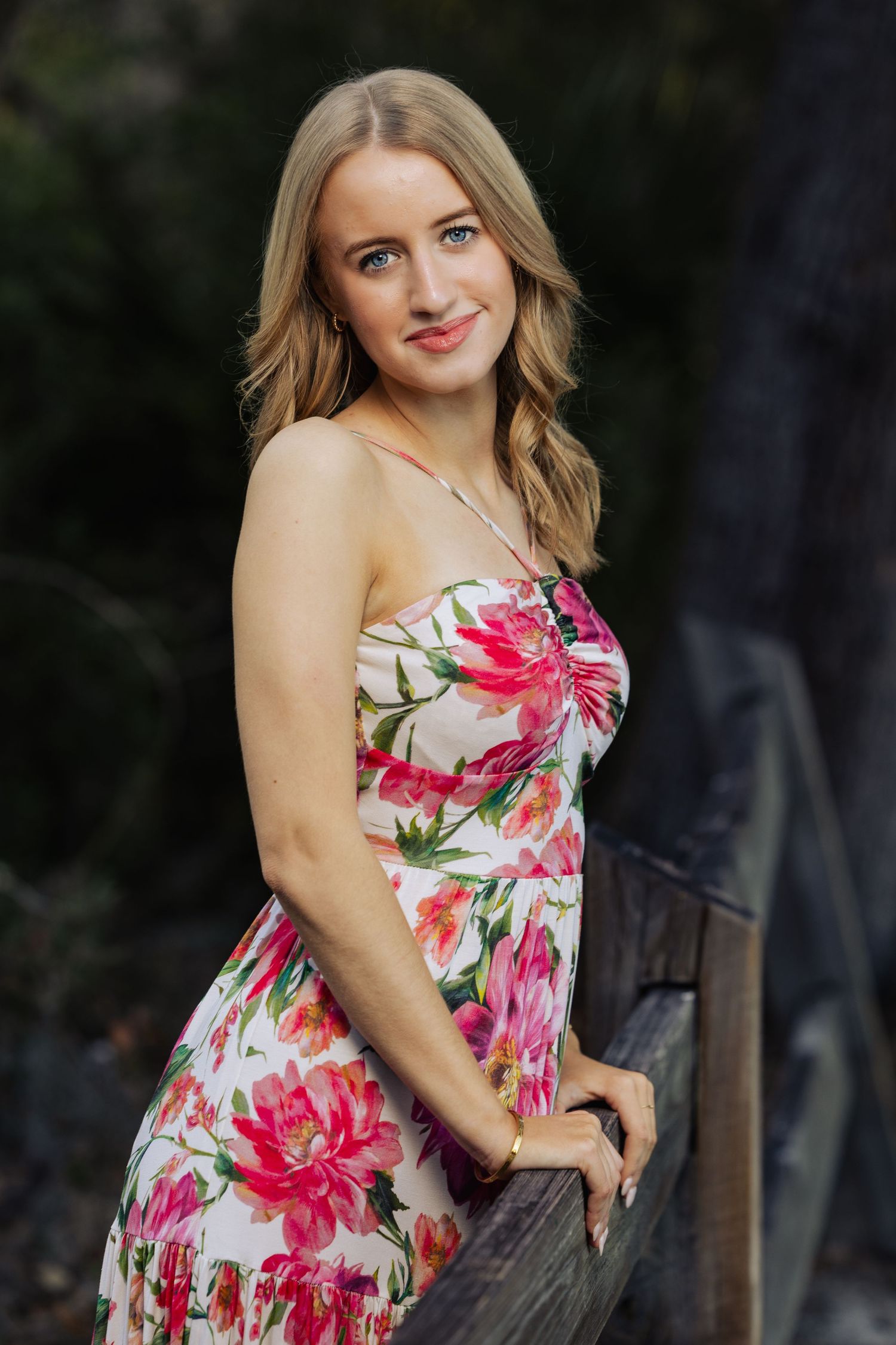 Young blonde woman in pink floral sundress leaning on wooden fence outdoors.