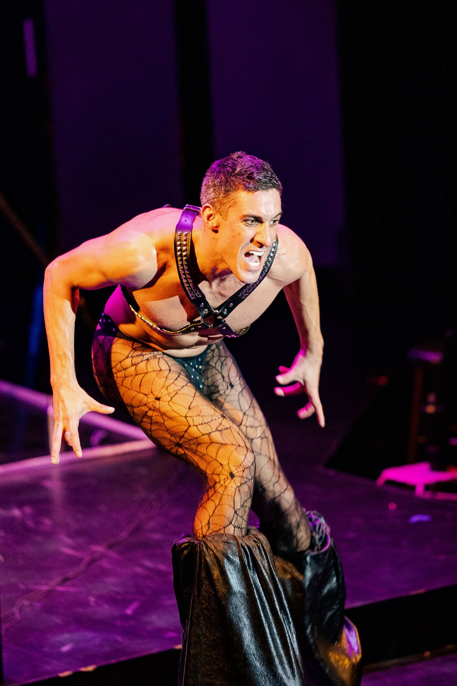 Intense male burlesque performer in fishnet tights and leather harness snarls at audience on purple-lit stage.