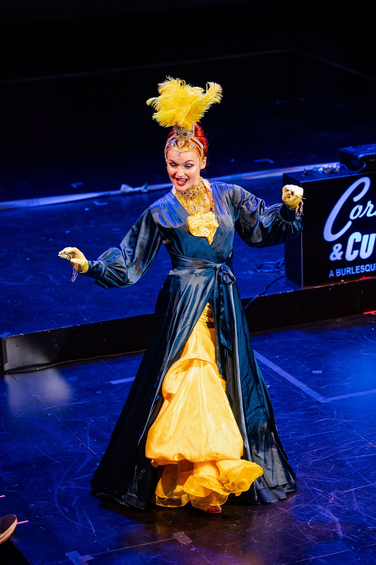 Burlesque dancer in elaborate blue and yellow gown with feathered headdress performing on stage.