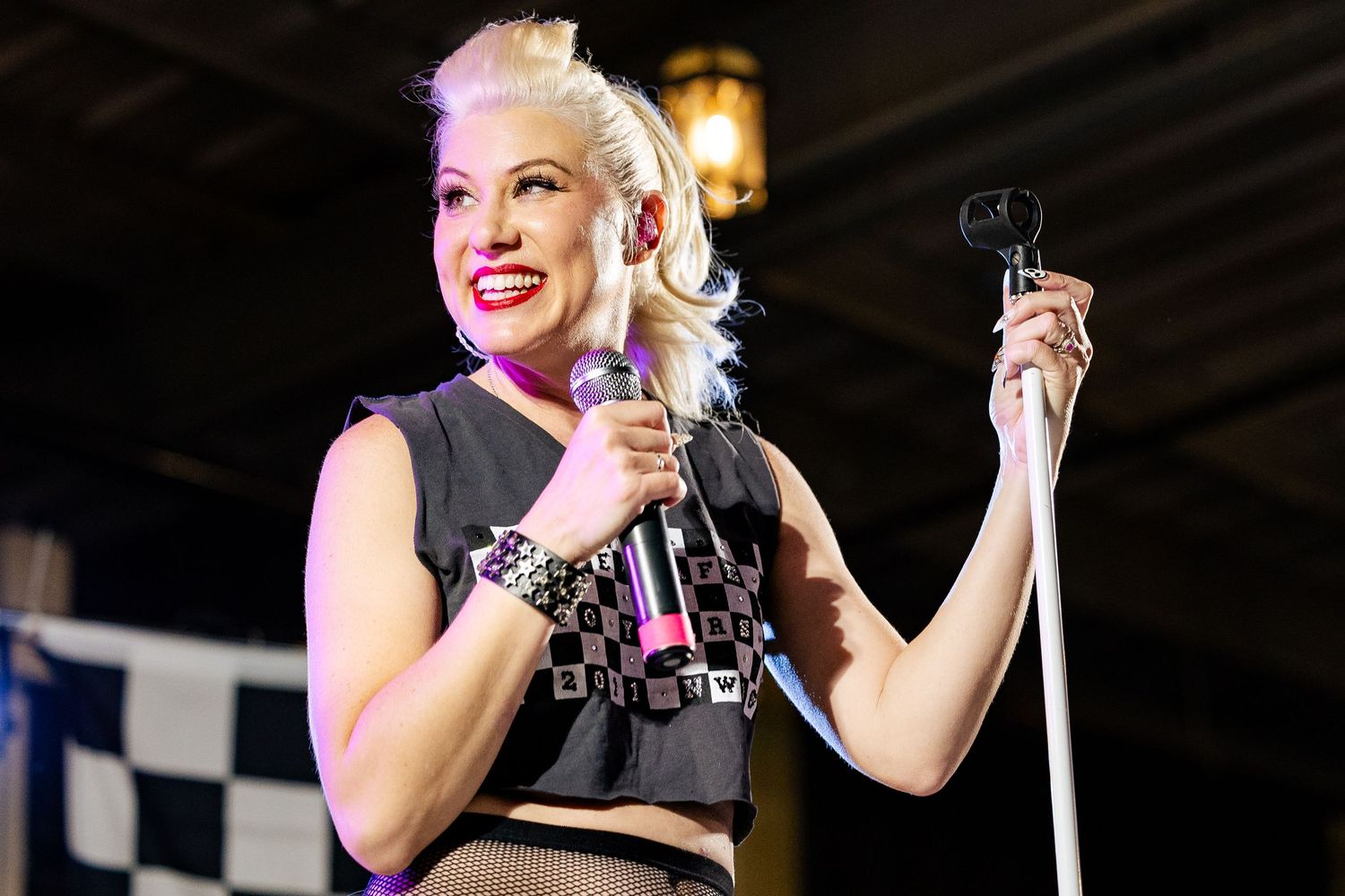 Blonde female vocalist smiles on stage holding microphone stand at live concert.