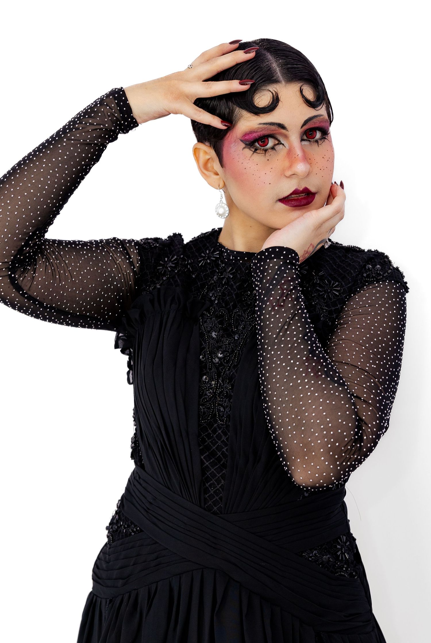 A model in a black sheer dotted dress poses with dramatic gothic makeup and styled hair.