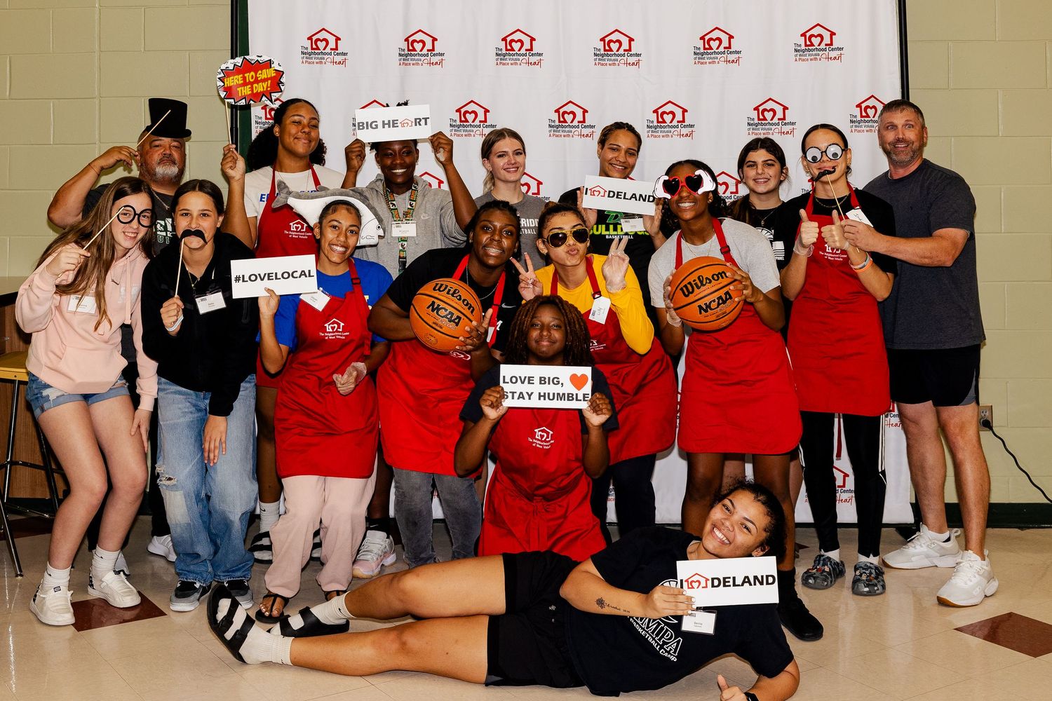 Energetic group of volunteers holding basketballs and props in front of a DeLand branded banner.
