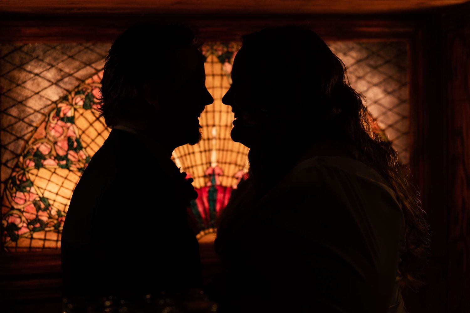 Romantic silhouette of bride and groom facing each other before glowing stained glass fireplace.