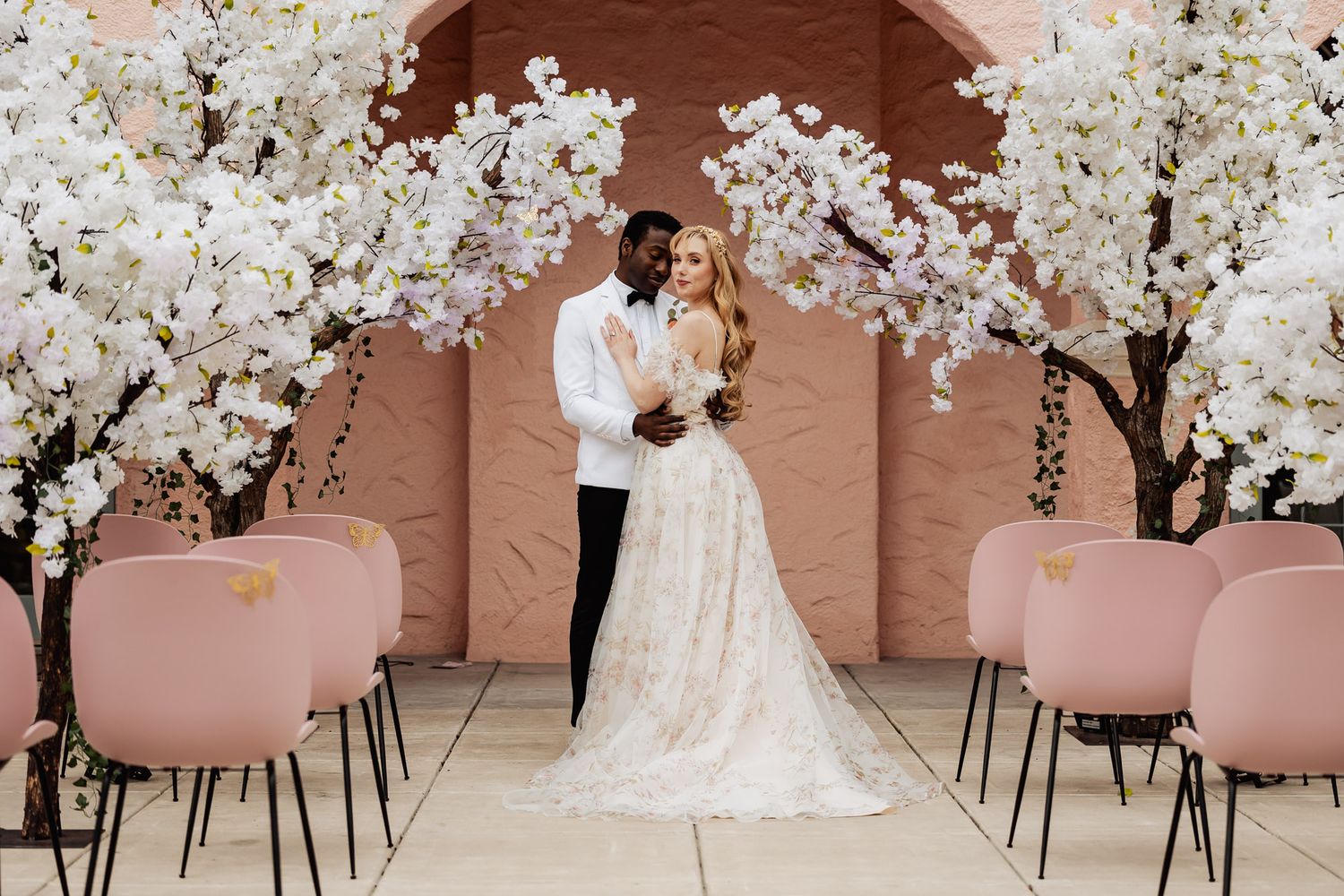 Interracial wedding couple standing at altar between white cherry blossom trees with pink chairs.