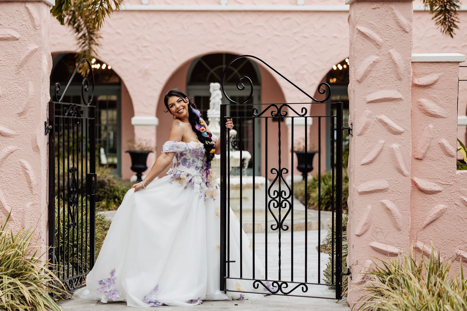 Bride in white floral off-shoulder gown holds champagne flute while posing at black iron gate.