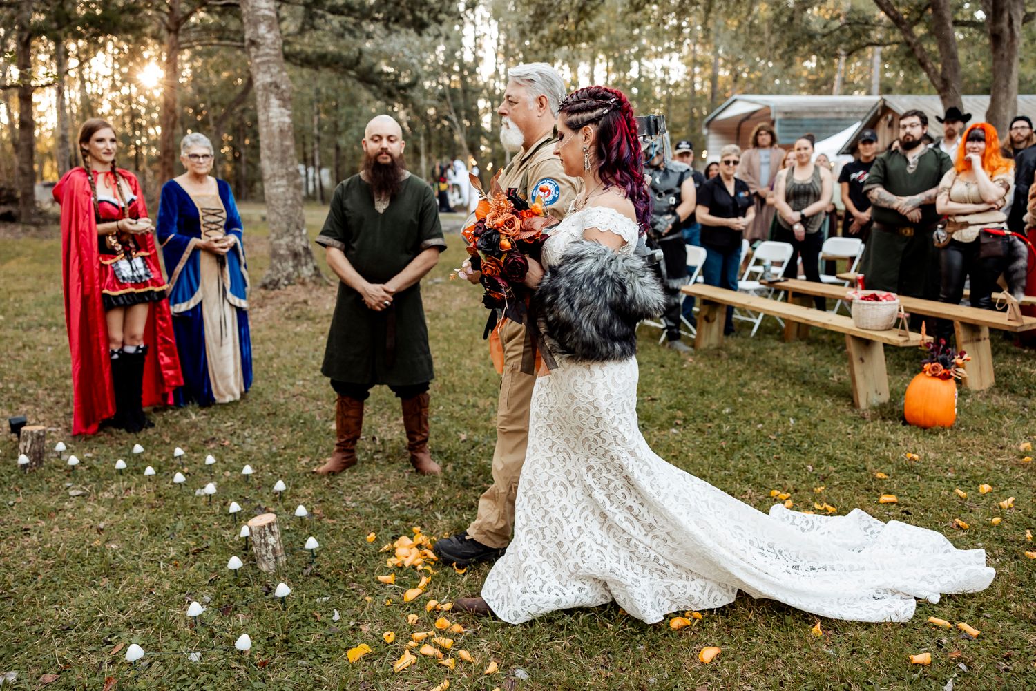 Bride in lace gown with colorful bouquet walks down outdoor aisle at Halloween-themed wedding.