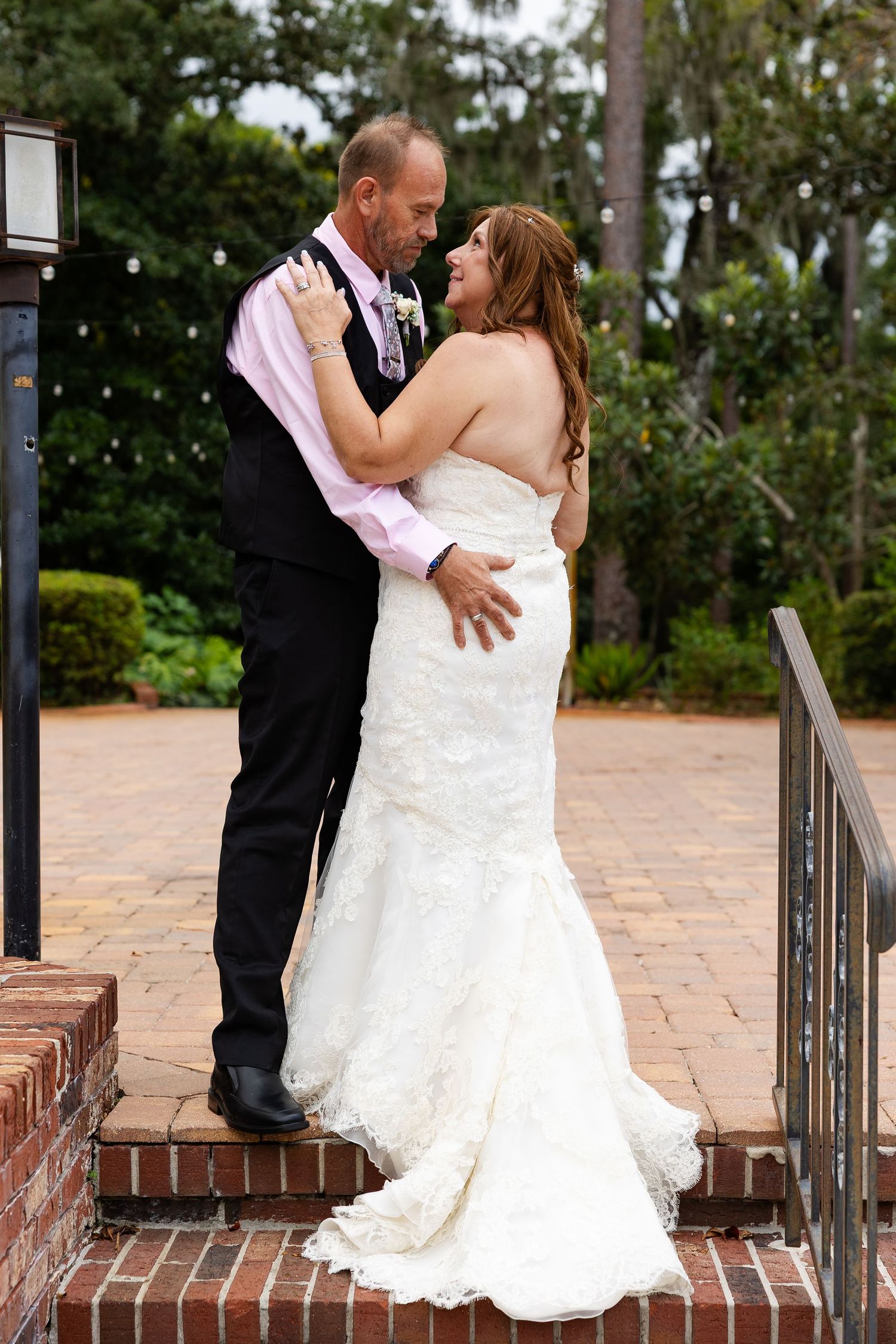 Bride and groom share an intimate embrace on brick steps surrounded by lush greenery at outdoor wedding venue.