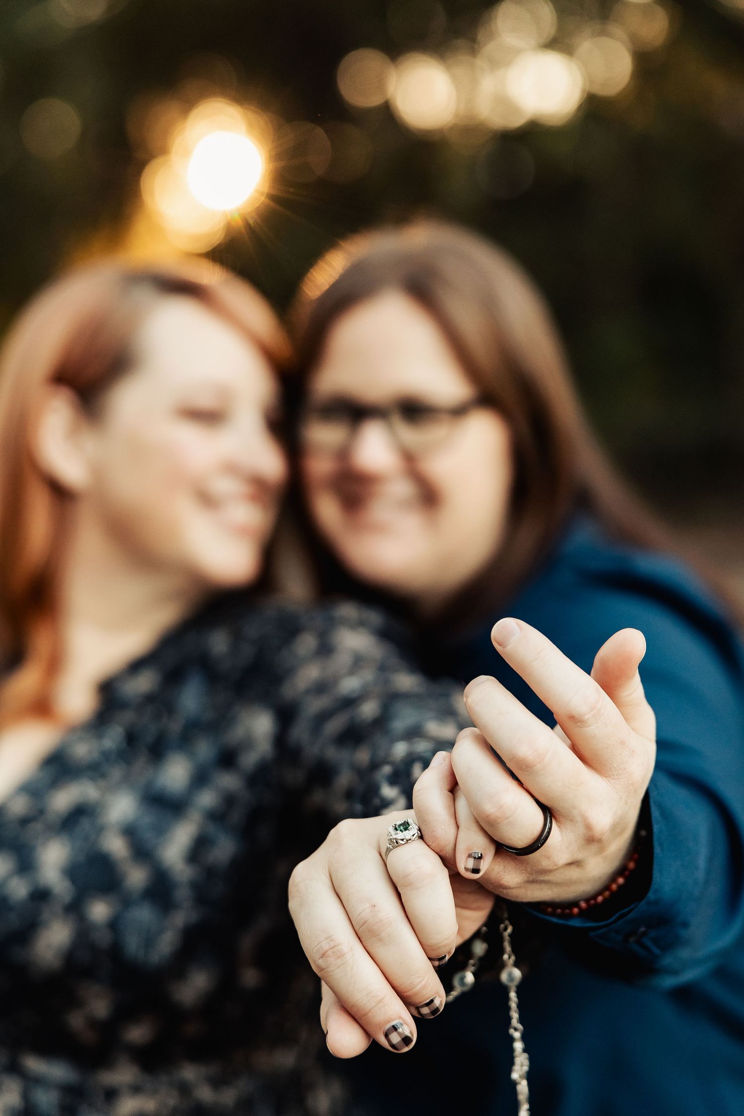 Engaged couple displays their rings close to camera, smiling softly with warm bokeh lights in the background.