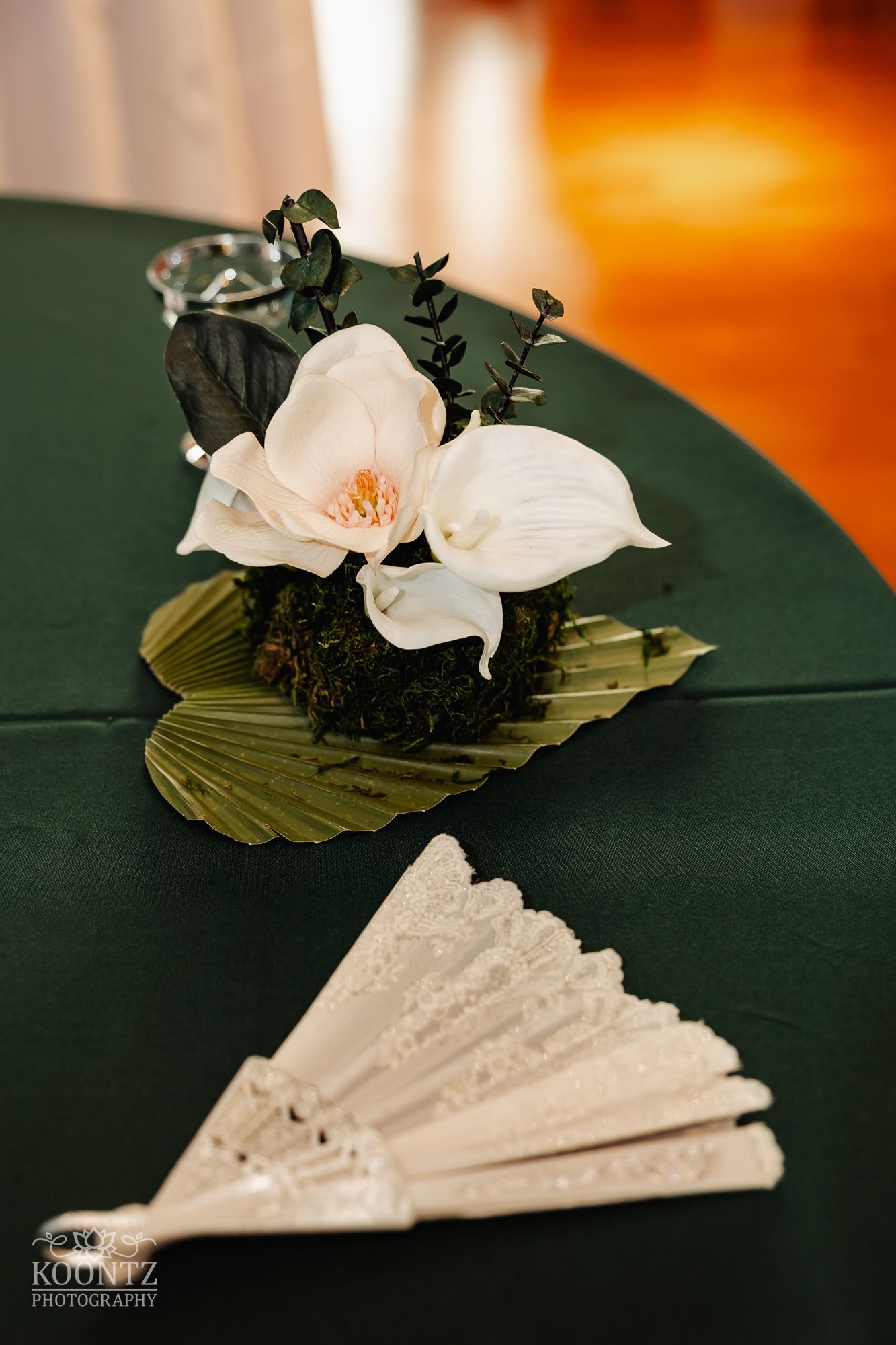 White orchid flower arrangement with greenery on dark surface next to decorative fan.