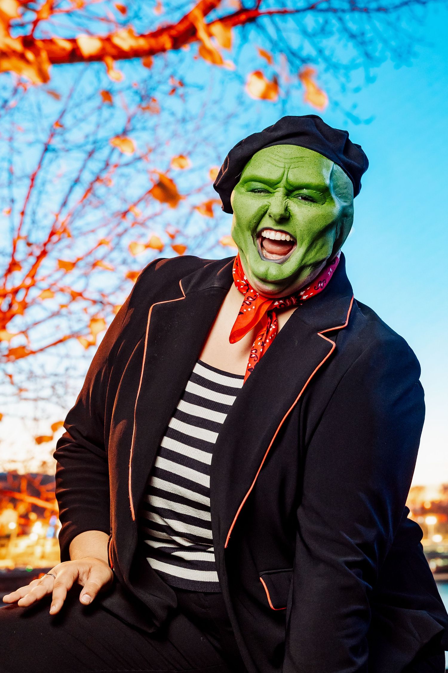 Theatrical green-faced performer in beret and striped shirt against autumn trees and blue sky.