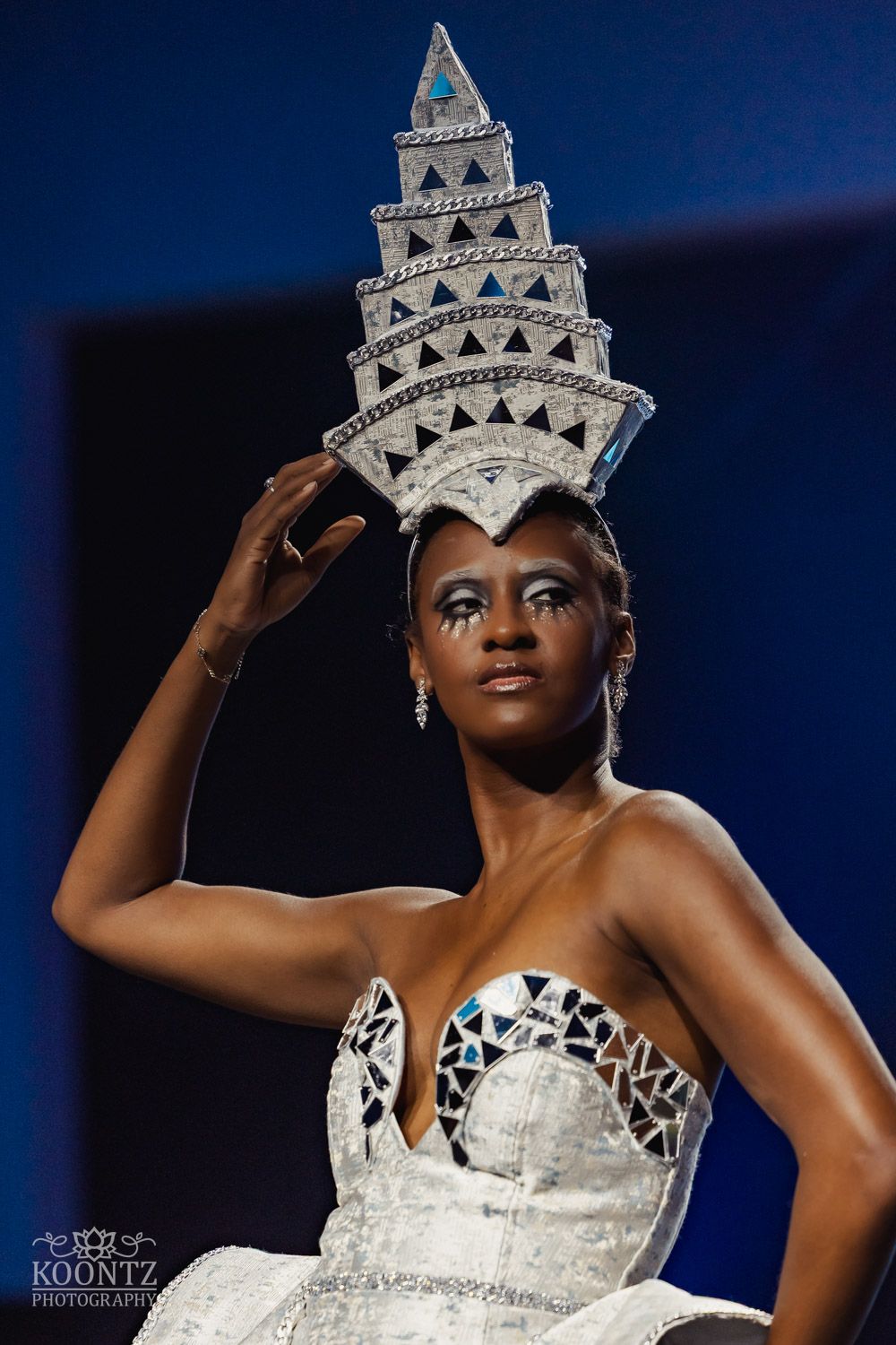 Elegant pageant contestant in white strapless gown and ornate crown poses gracefully against dark background.