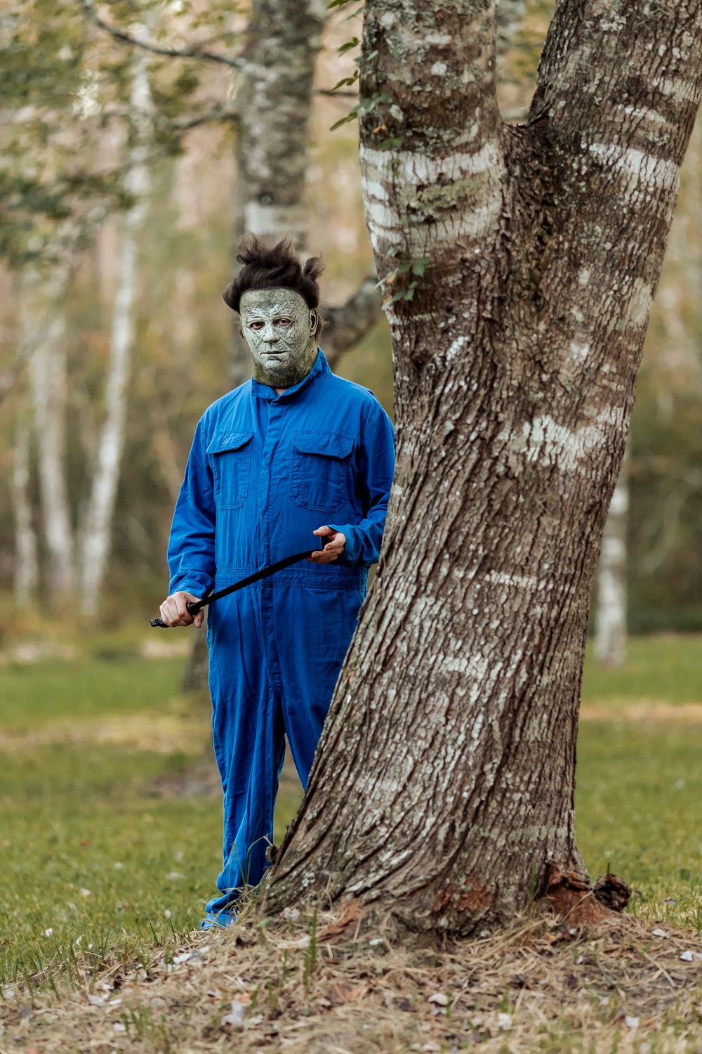 Figure in blue coveralls and horror mask standing against birch tree in autumn setting.