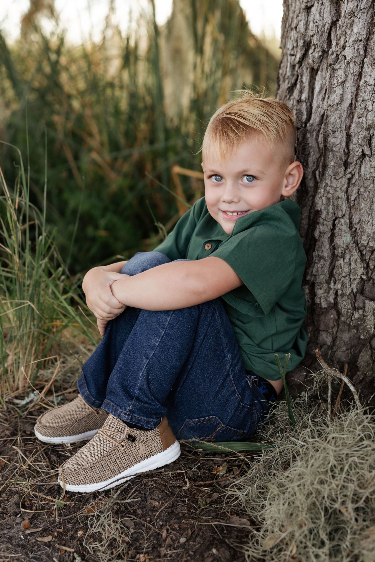 Child in green shirt sits casually against tree trunk outdoors.