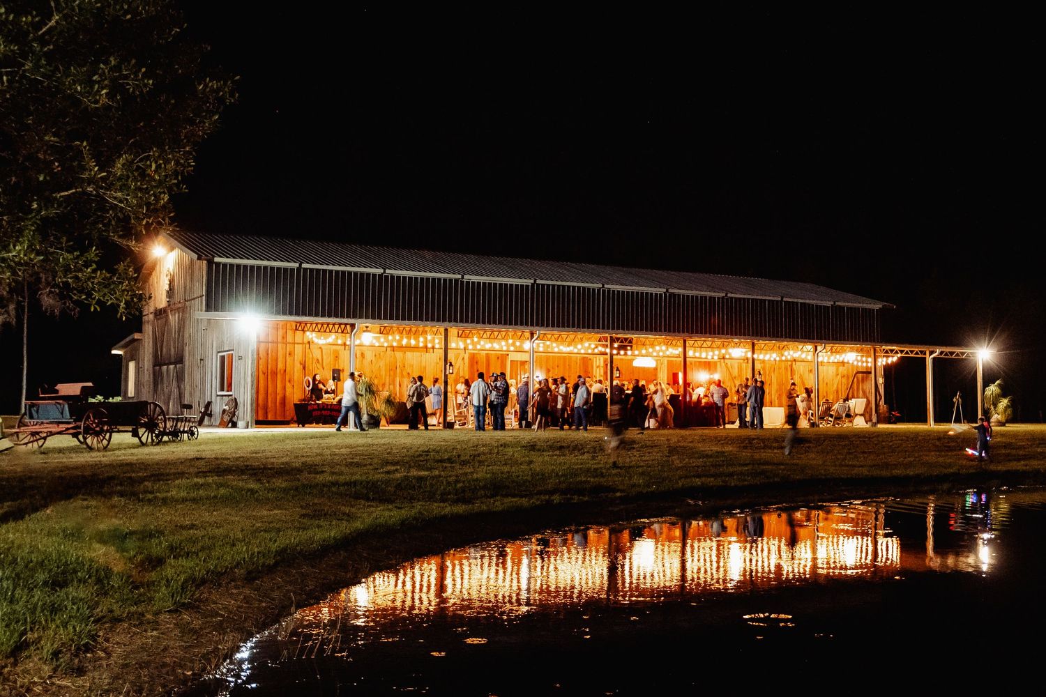 Barn wedding venue lit up at night with reflection in nearby pond.