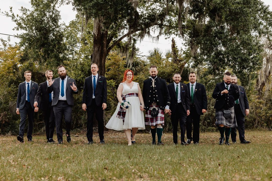 Wedding party poses outdoors with bride in white dress and groom in Scottish tartan kilt among moss-draped trees.