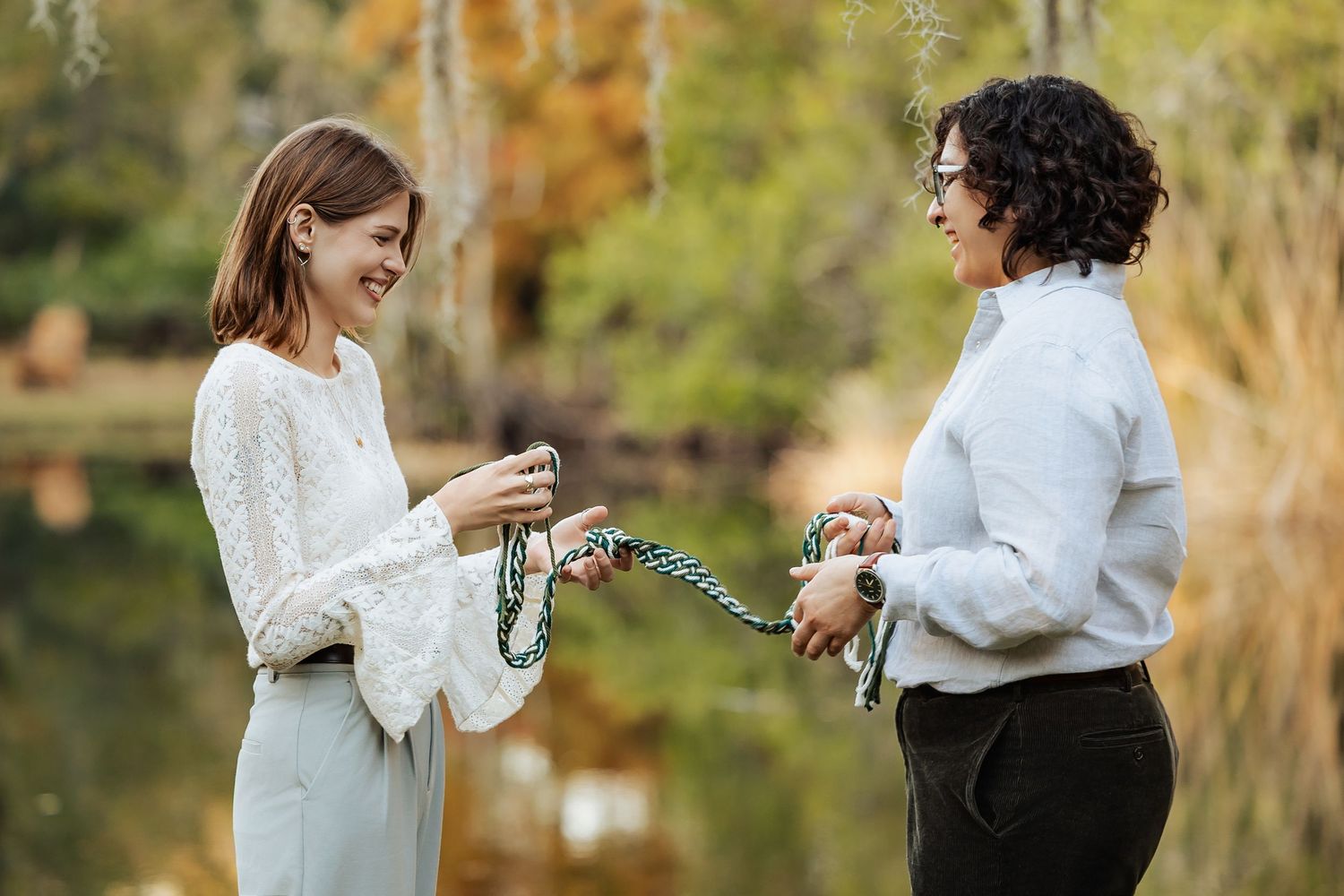 Two women laugh and exchange colorful beaded necklaces outdoors near a reflective pond in autumn.