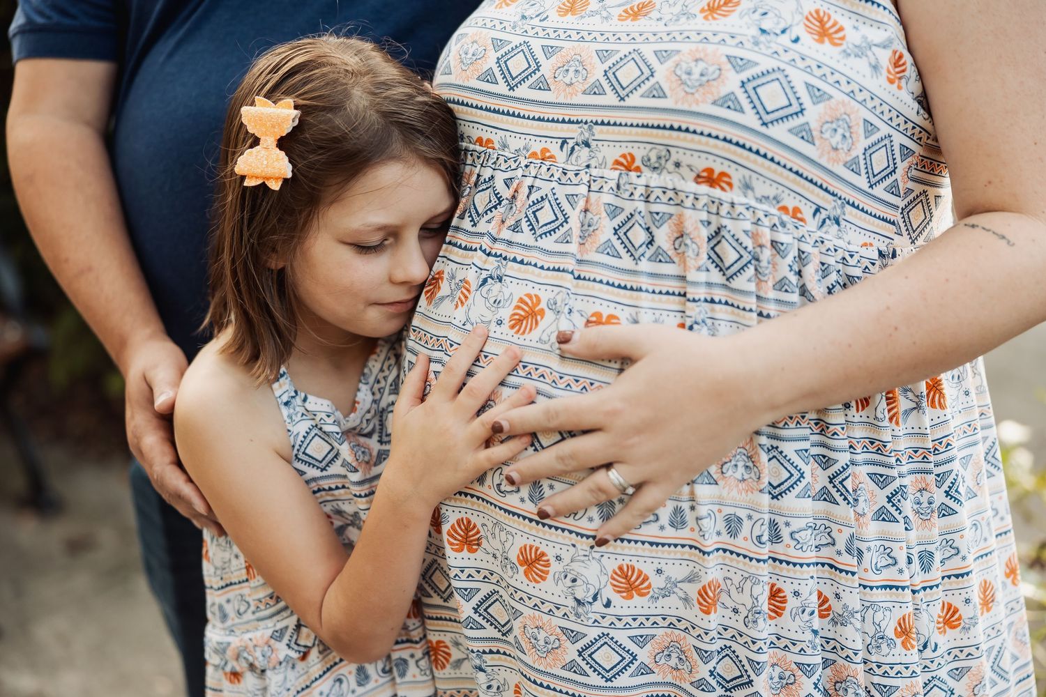 A young girl in a floral dress hugs a pregnant woman's belly tenderly outdoors.