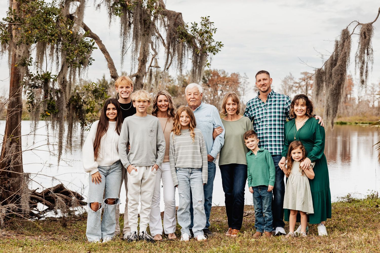 A large multigenerational family poses together outdoors near a moss-draped tree by a lake.