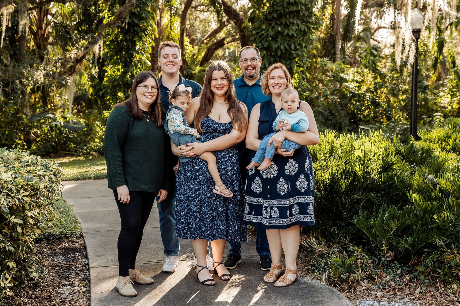 A multigenerational family of seven smiles together on a sunlit garden pathway.
