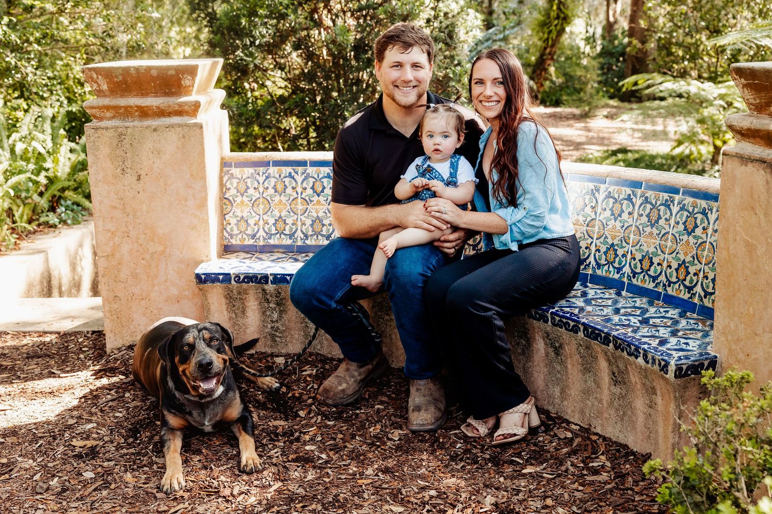 Family of three with a Rottweiler dog sits on a decorative tiled bench in a lush garden.